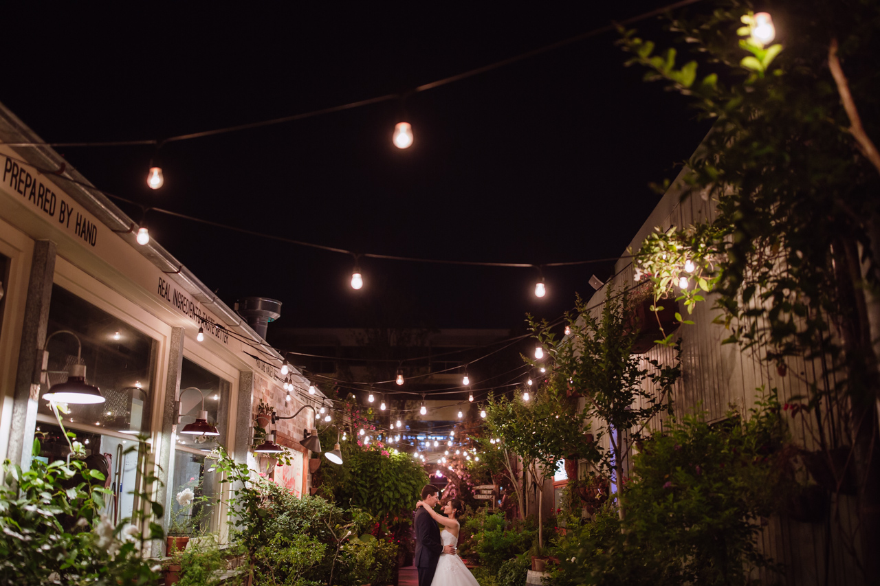 A man and woman in wedding attire, embracing in an open corridor with fairy lights