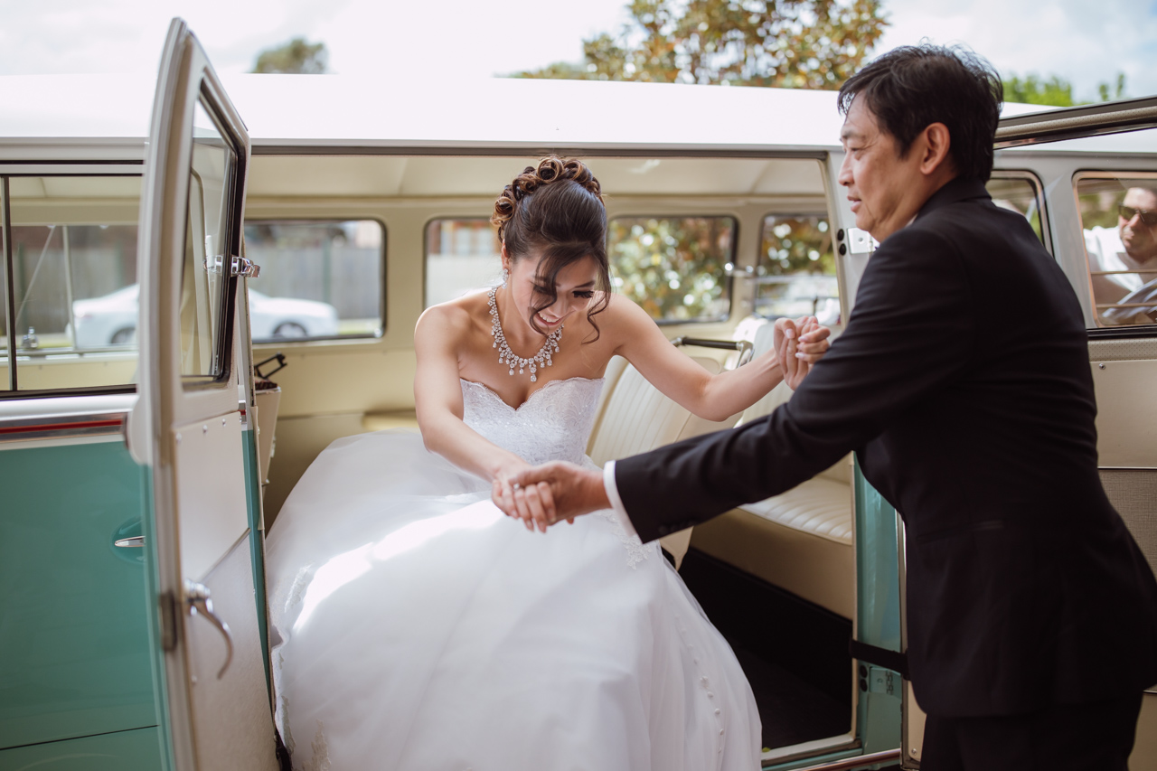 A woman in a wedding dress being helped out of a kombi camper van by her father