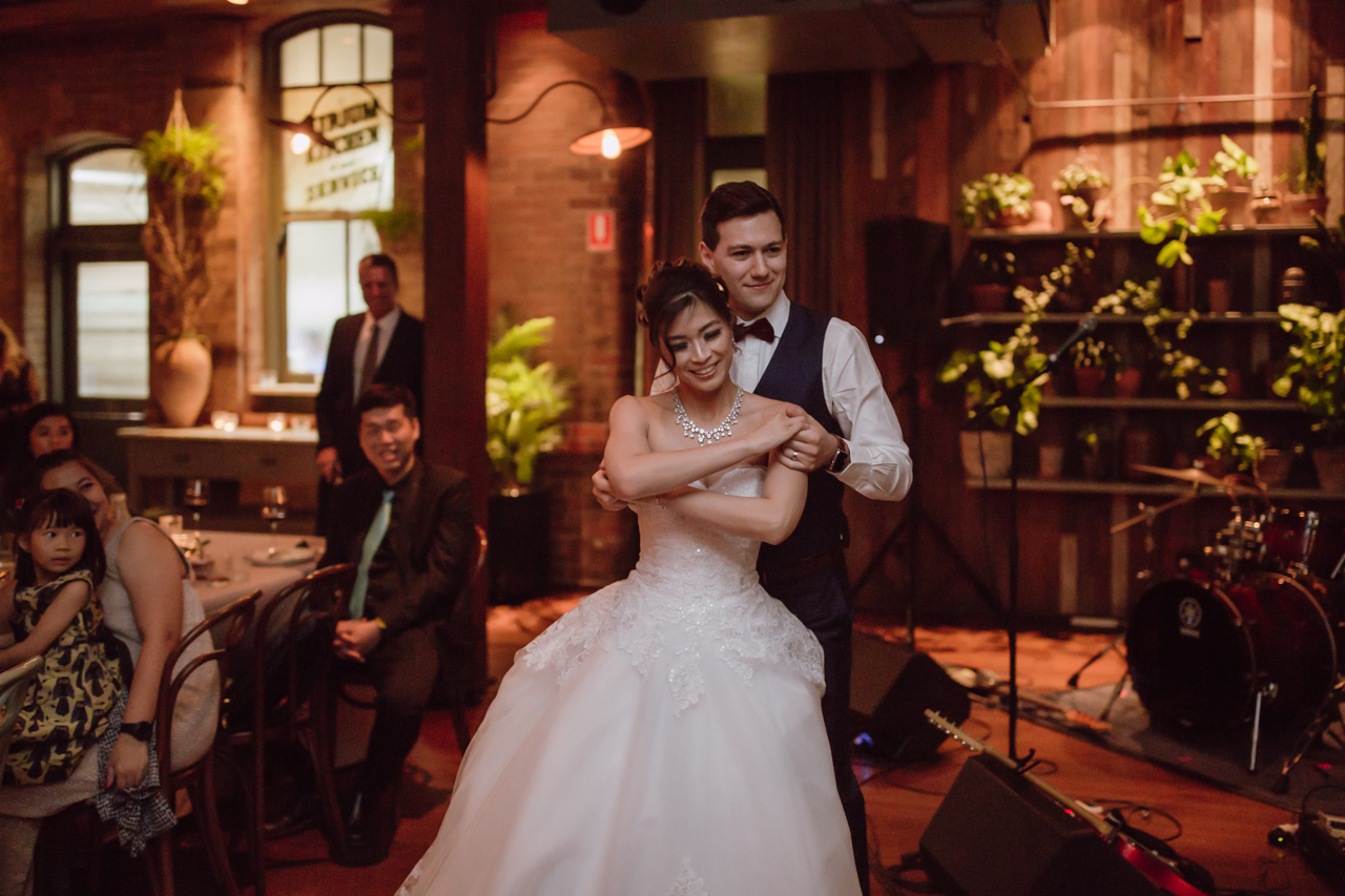 A married couple dancing in a room. Some wedding guests look on from the side. There are some musical instruments and microphones set up in the background