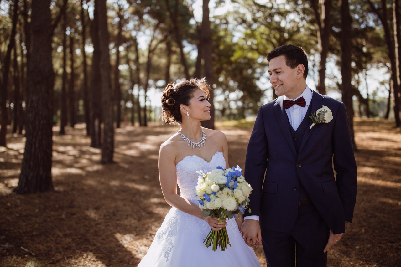 A bride and groom looking at each other and holding hands. The background mostly consists of trees