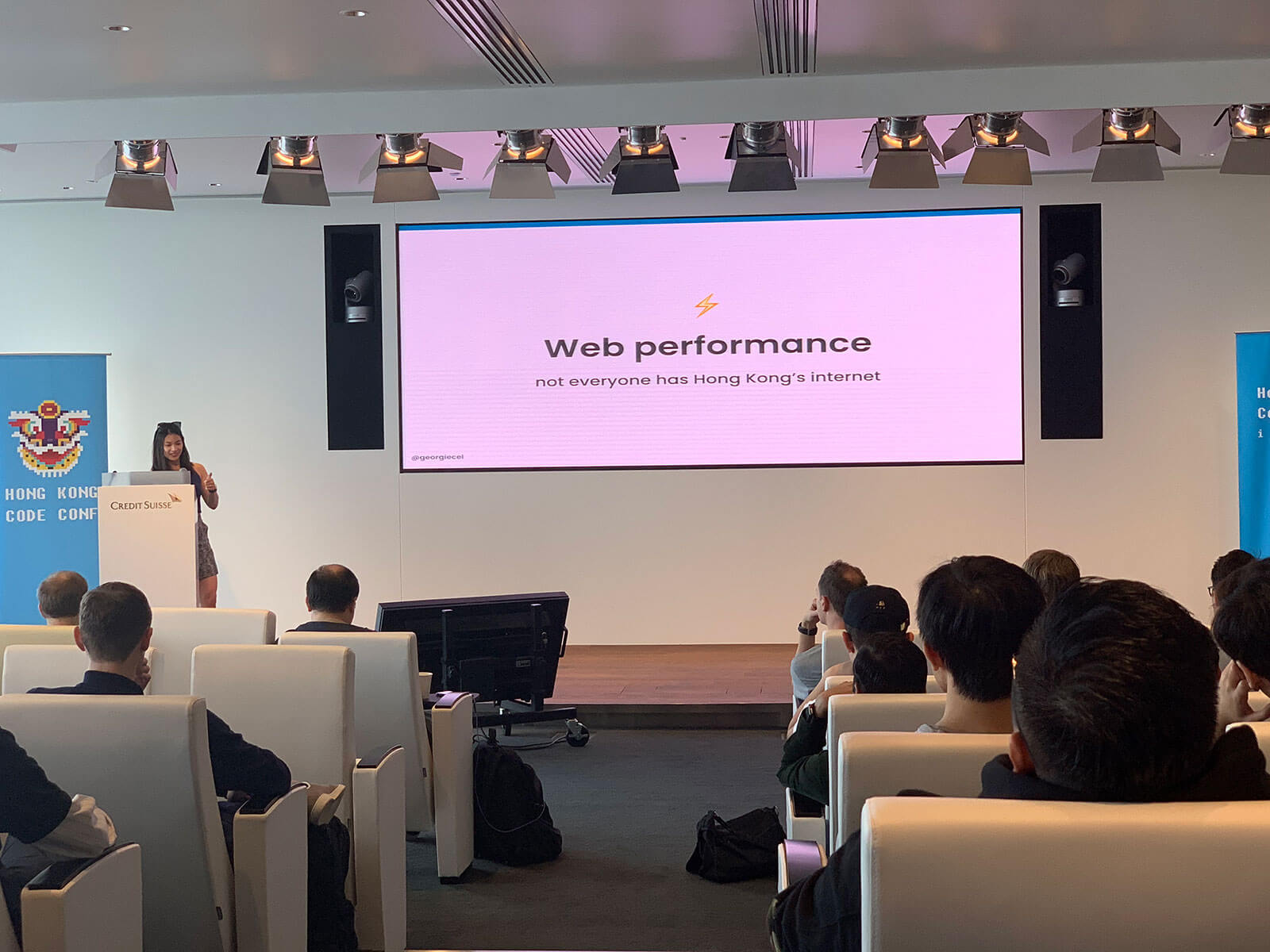 A conference room with white chairs with people seated in them. At the front of the room on a stage is a woman standing at a podium. The screen backdrop on the stage reads “Web Performance”