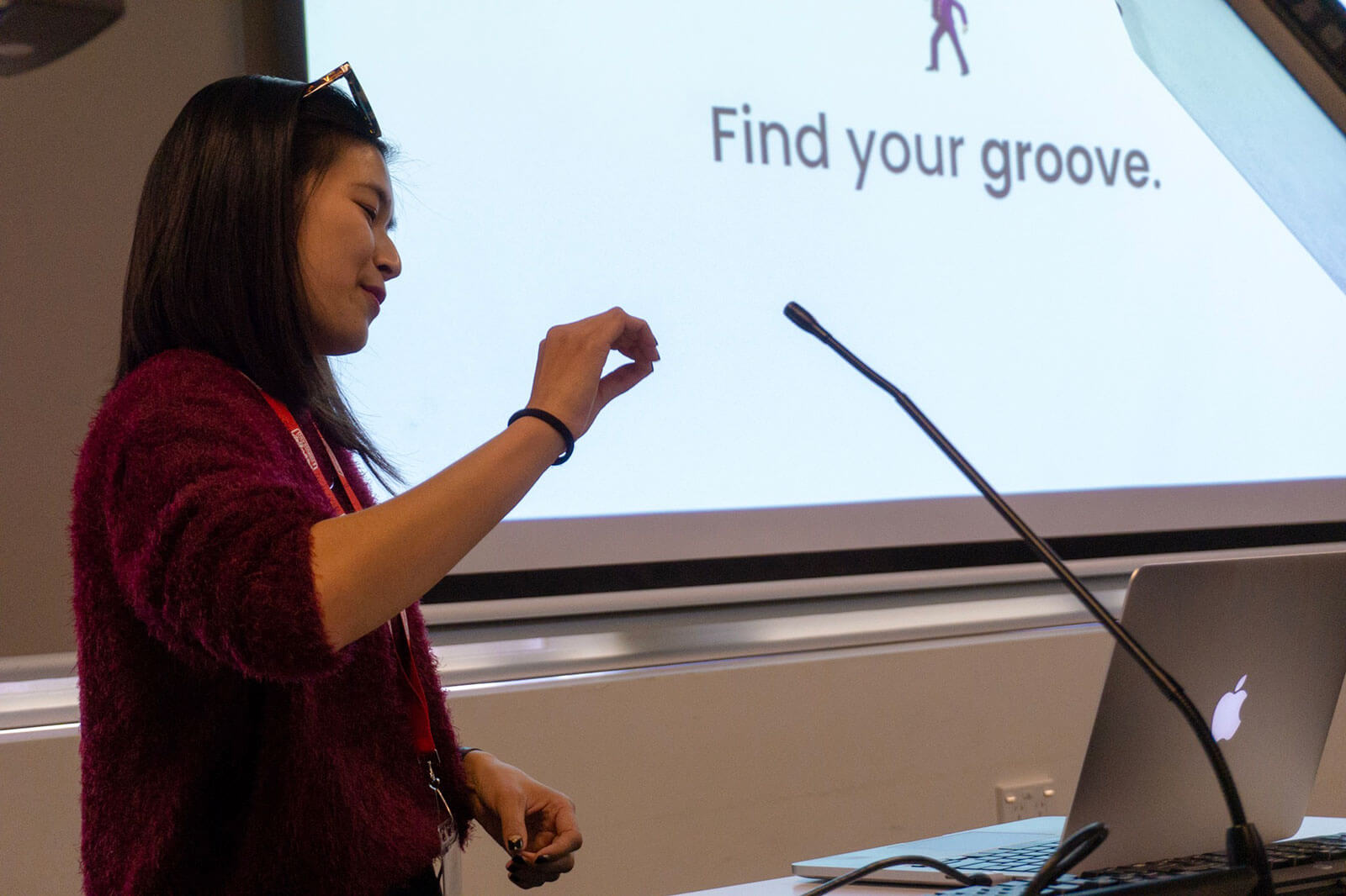 A woman in a red sweater in front of a screen reading “Find your groove”. The thumb and fingers of one of her hands is closed together in an o shape