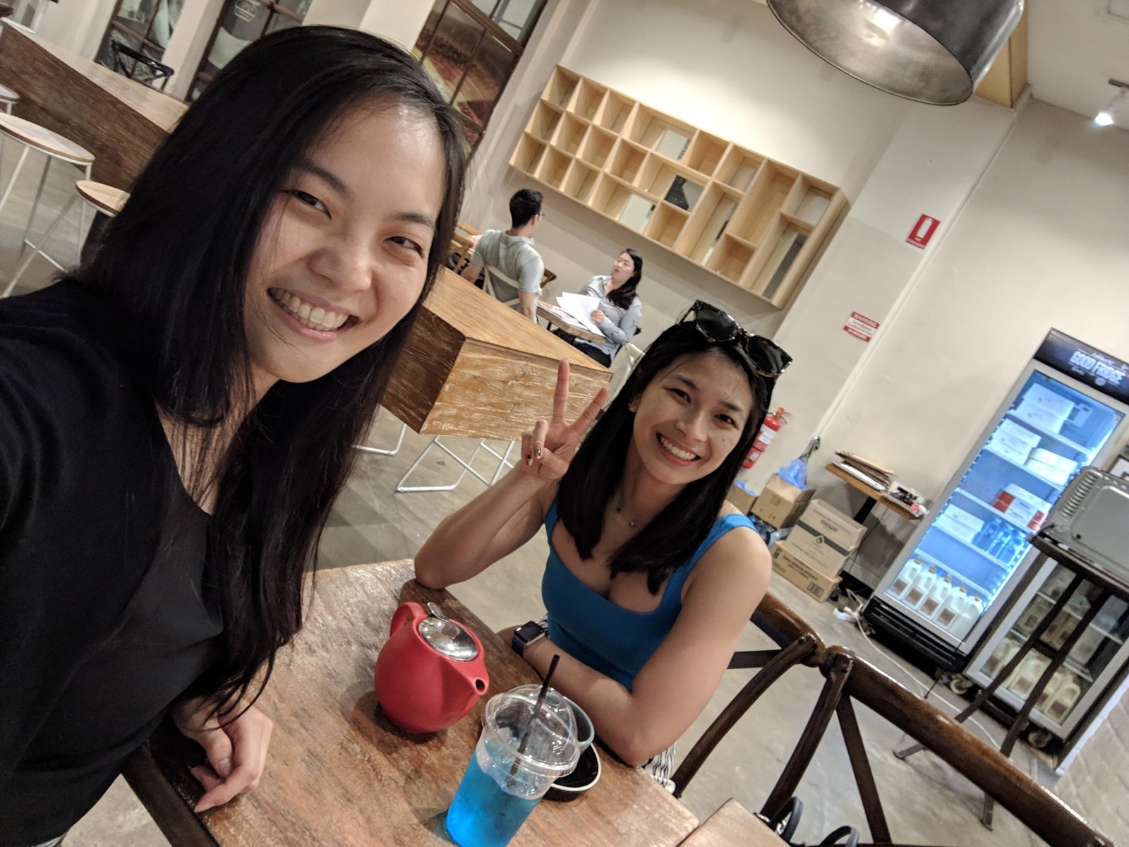 A selfie with two women in it; both are smiling. The woman taking the selfie is wearing black and the other woman is wearing blue. They are seated in a cafe and on the table is a pot of tea and a plastic cup of blue lemonade.