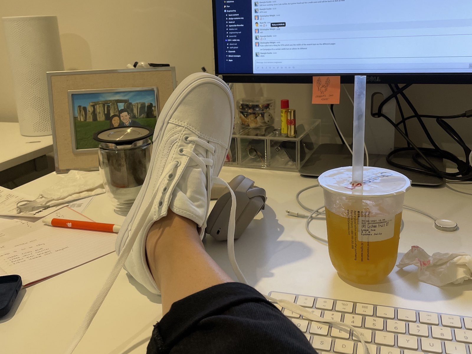 A woman’s foot with a half-laced white sneaker on it, propped up on a desk. In the background is a computer display and some other miscellaneous items.