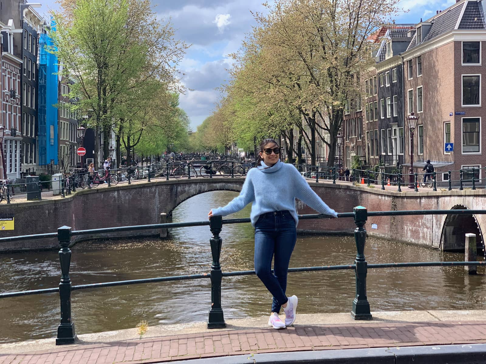 A woman in a light blue sweater leaning back against the railing of a bridge with a canal below it