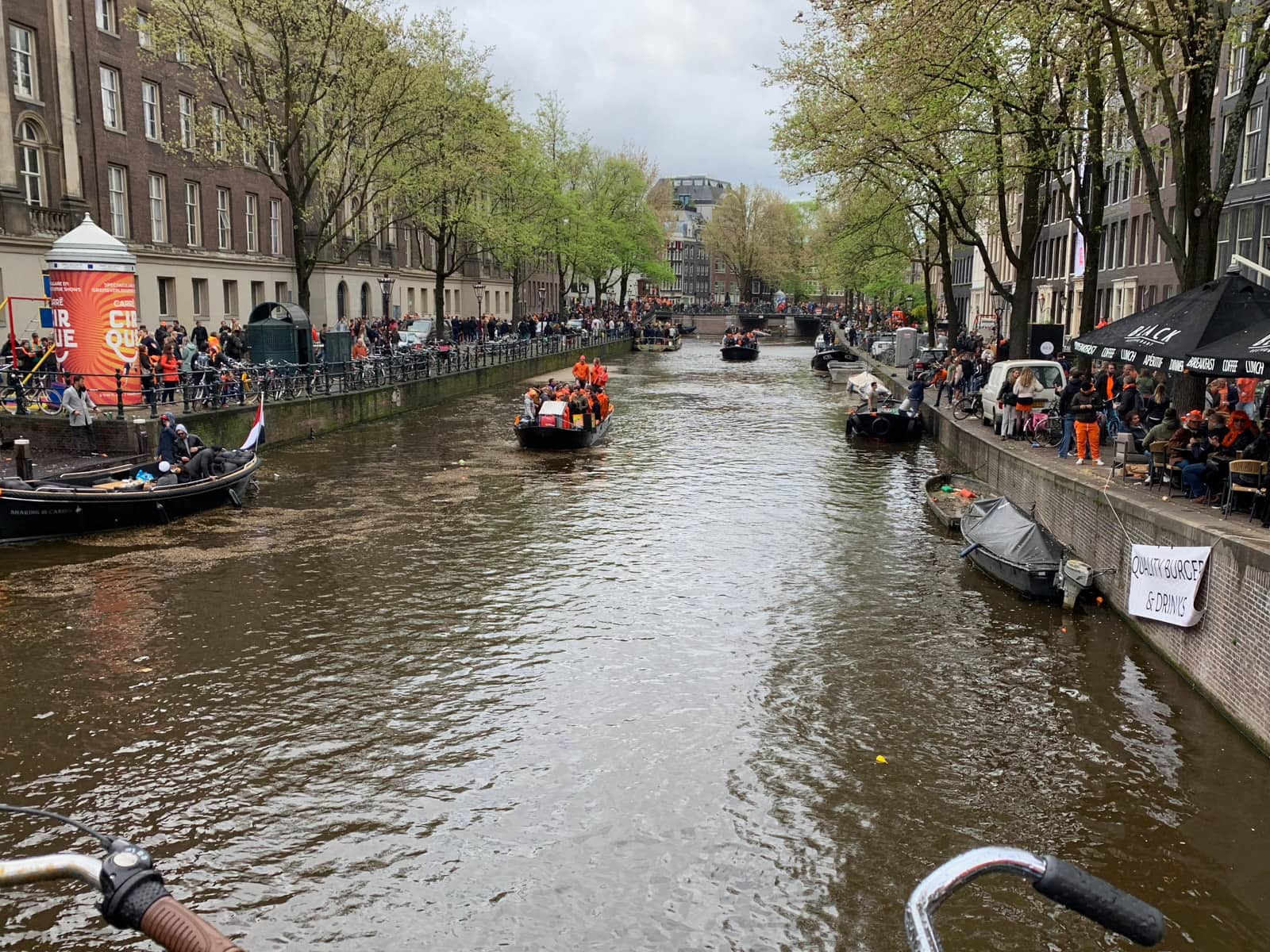 A canal river as seen from a perpendicular bridge. There are several bits of people wearing orange and celebrating, and the banks are also filled with people wearing orange