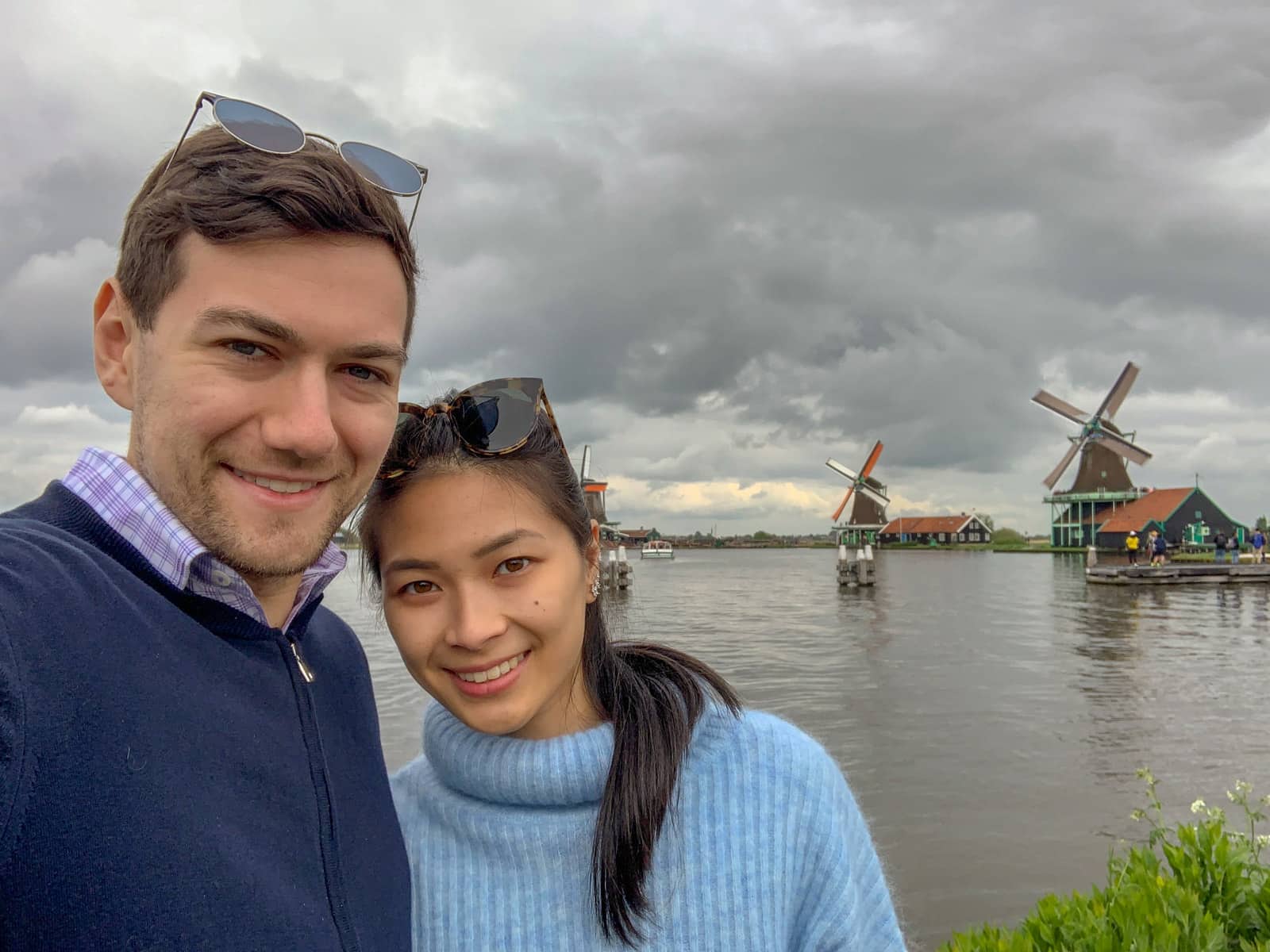 A man and woman taking a selfie in front of a river with Dutch windmills in the background. It is a very cloudy day