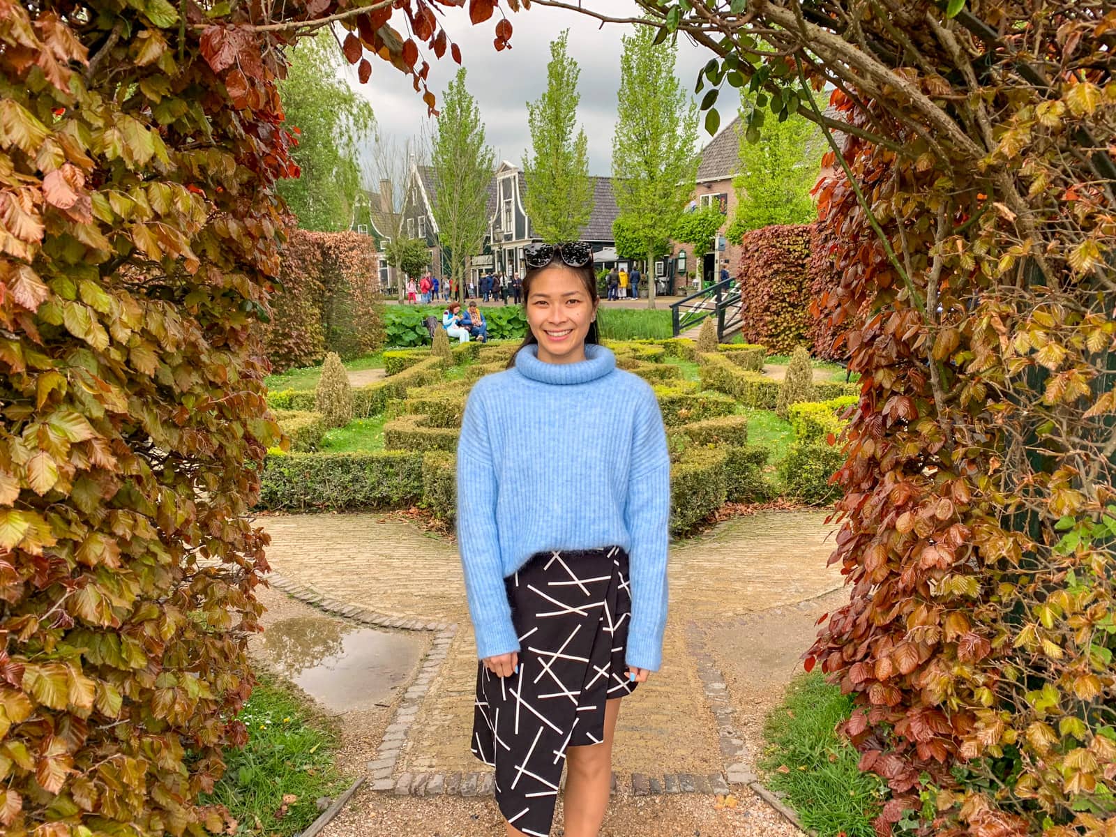 A woman in a blue sweater and black patterned skirt under an hedge arch of red and yellow coloured leaves. In the background is a small green garden maze.