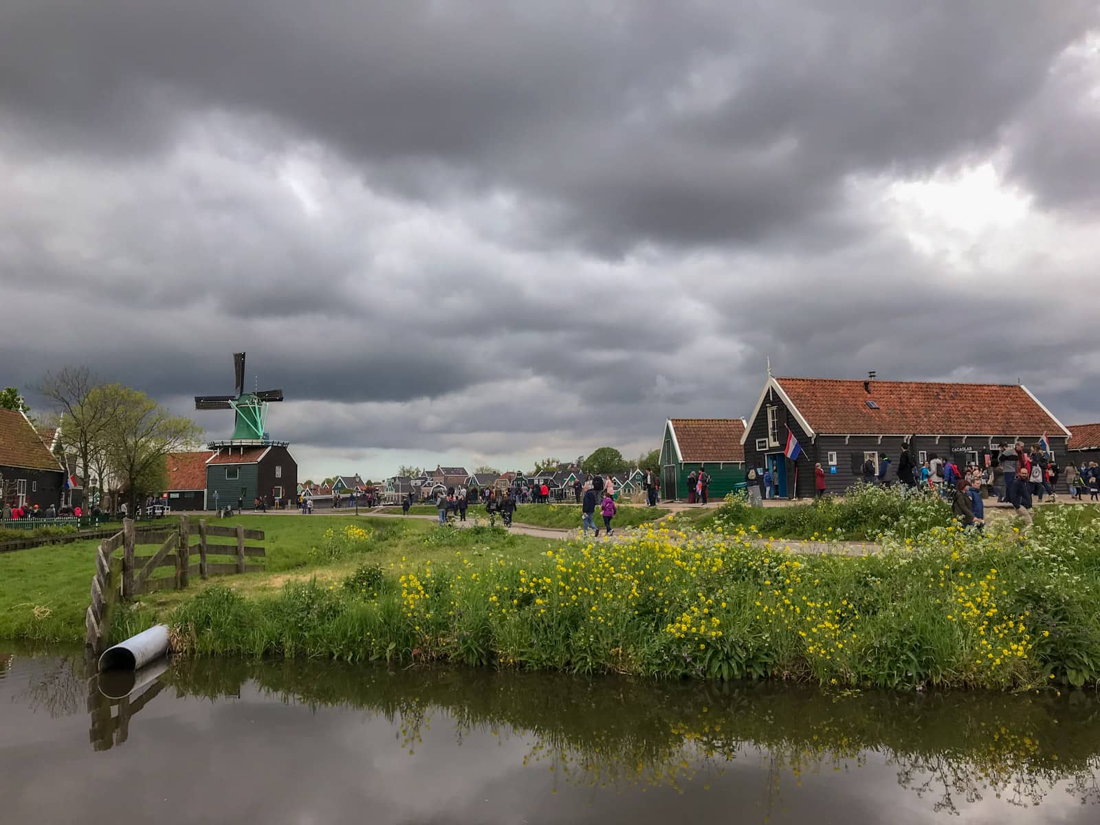 A small village with cottage houses and a windmill in the distance, as seen over a small dark-coloured moat of water. The sky is very cloudy and very grey.