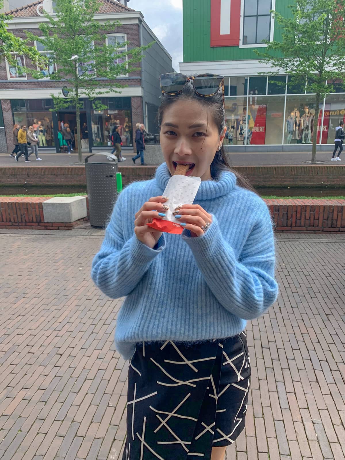 A woman in a blue sweater eating a deep-fried sausage out of a paper bag. In the background is an outdoor shopping mall
