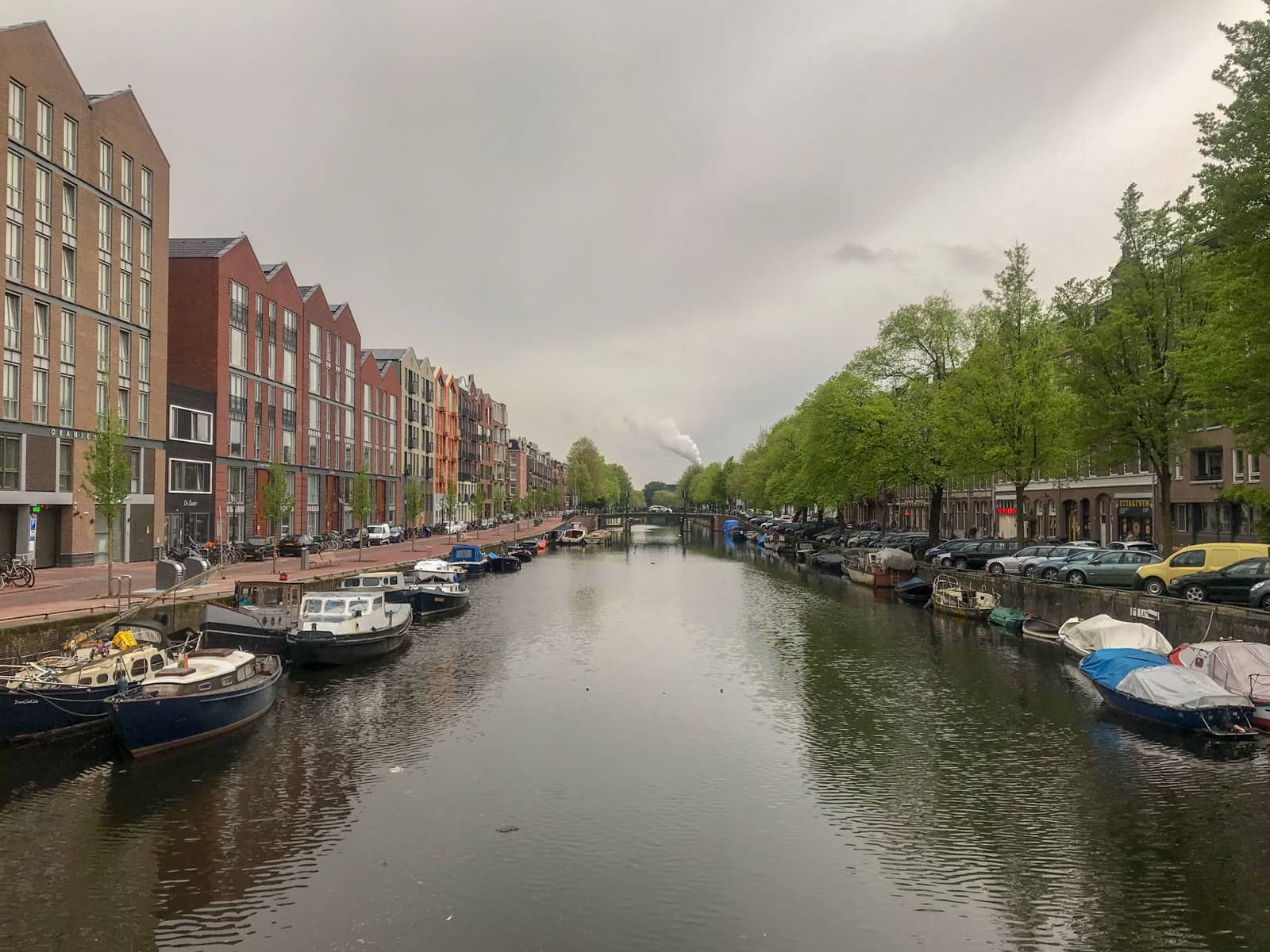 A canal in Amsterdam, seen from a bridge. The canal extends further away into the distance. Many canal boats are stationary on the sides of the canal.