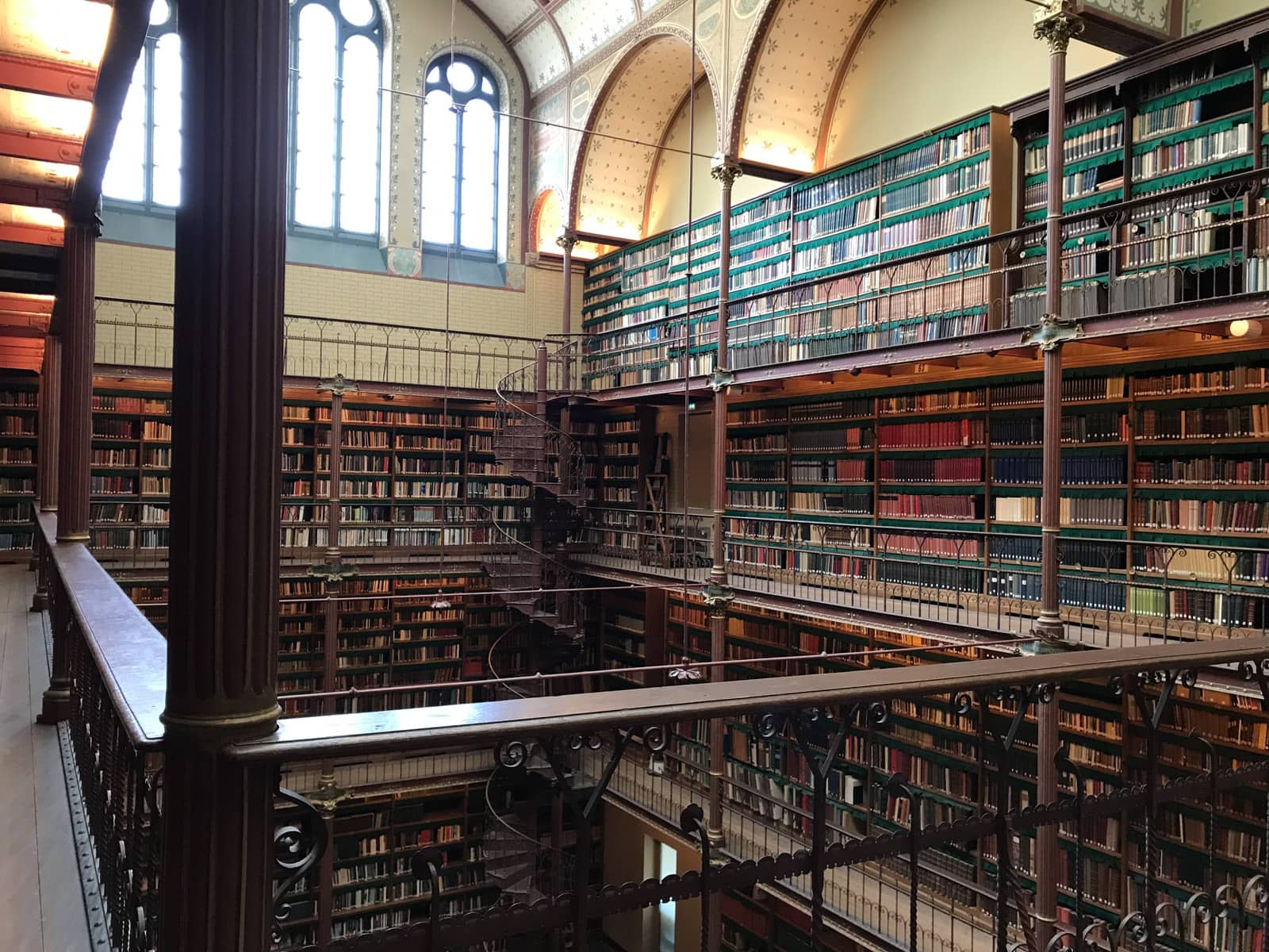 A historic-looking library seen from an indoor balcony. Many books line the walls and the decor is brown and antique. The books line many shelves all the way down the walls