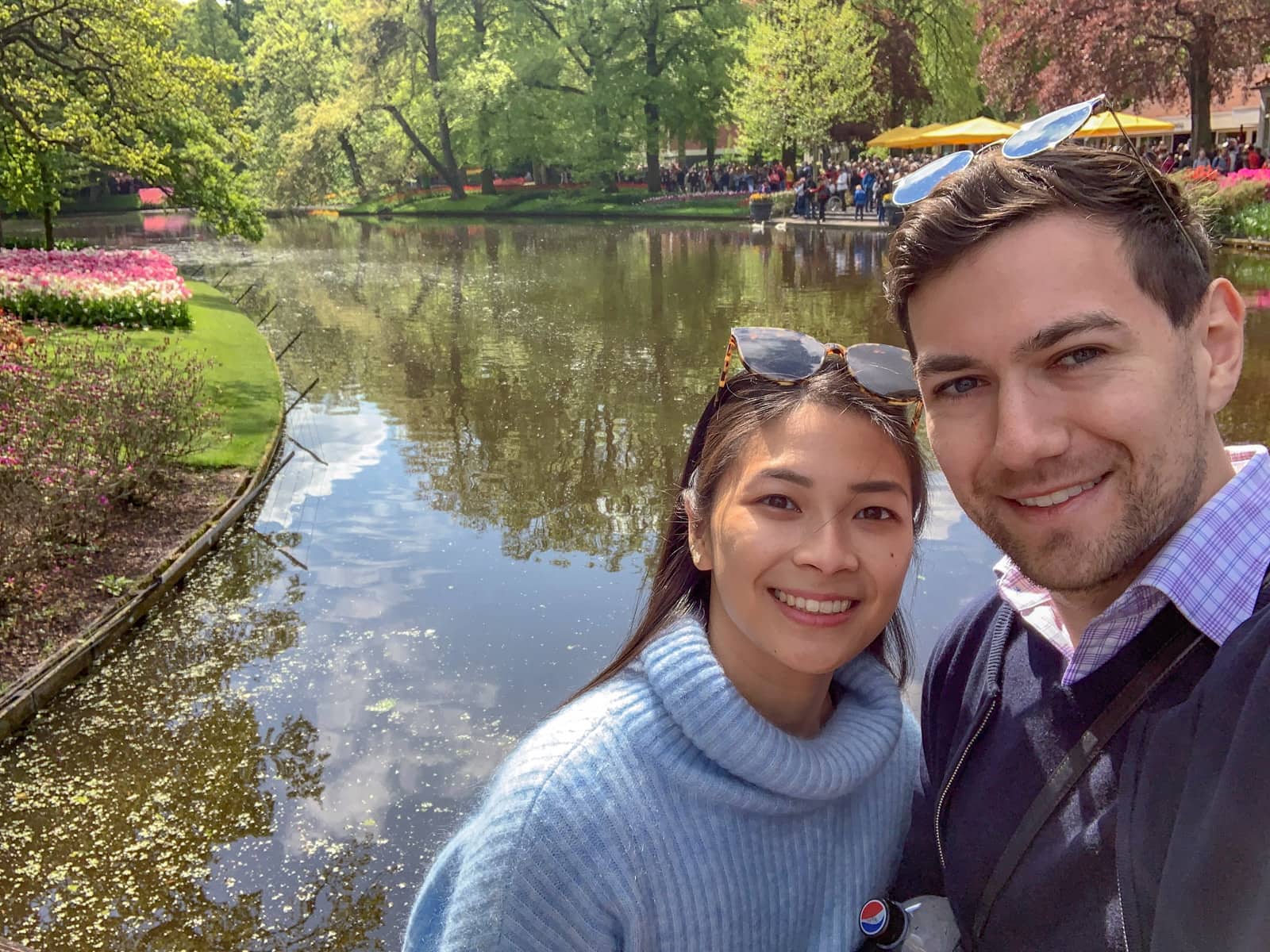 A man and woman smiling and taking a selfie in front of a river with several leafy debris in it. On the banks of the water are rows of planted tulips in bright colours.