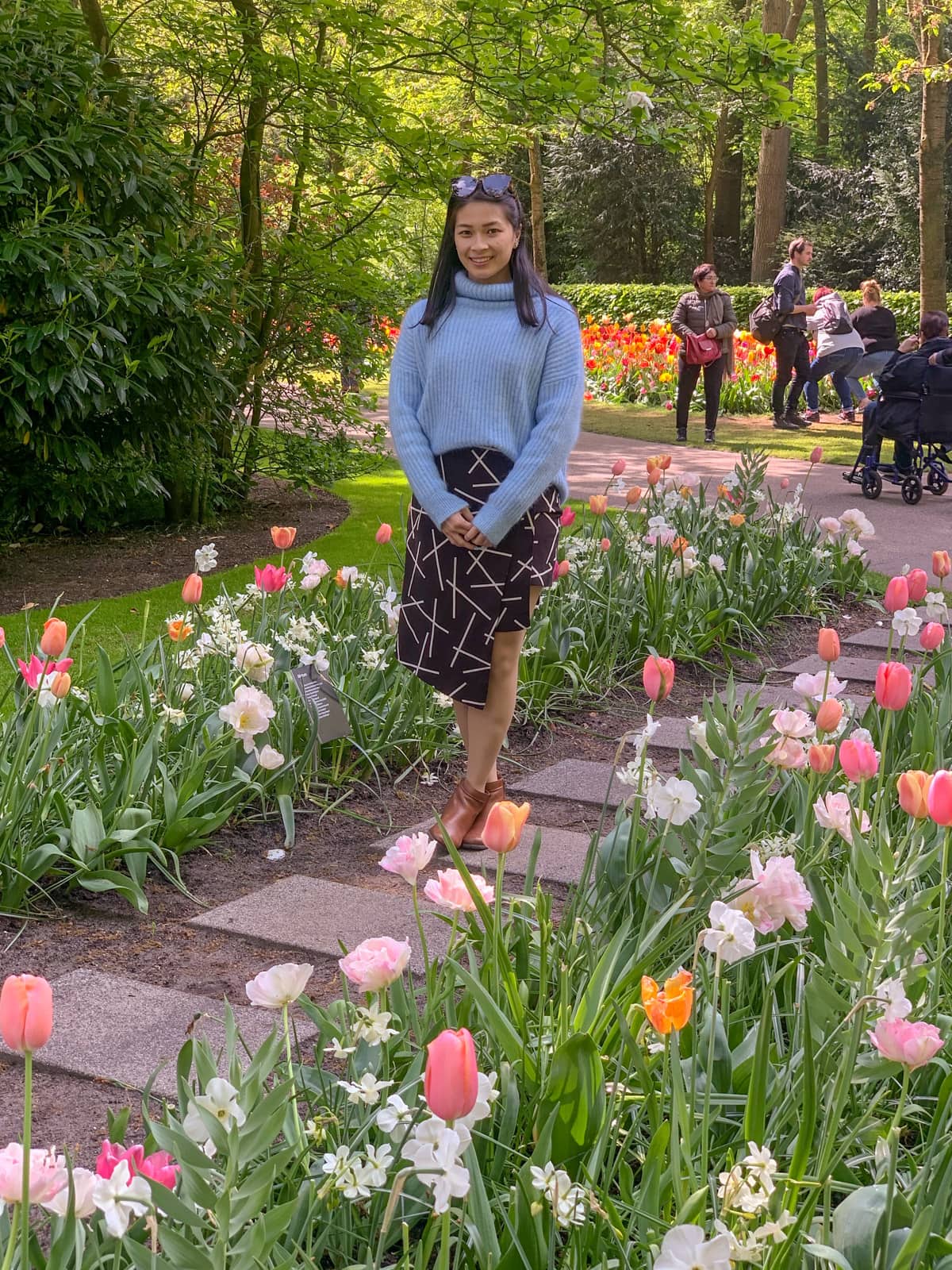 A woman in a light blue sweater and black patterned skirt standing on a square-paved stone path between two rows of tulips that are all pastel pink, orange, and yellow in colour