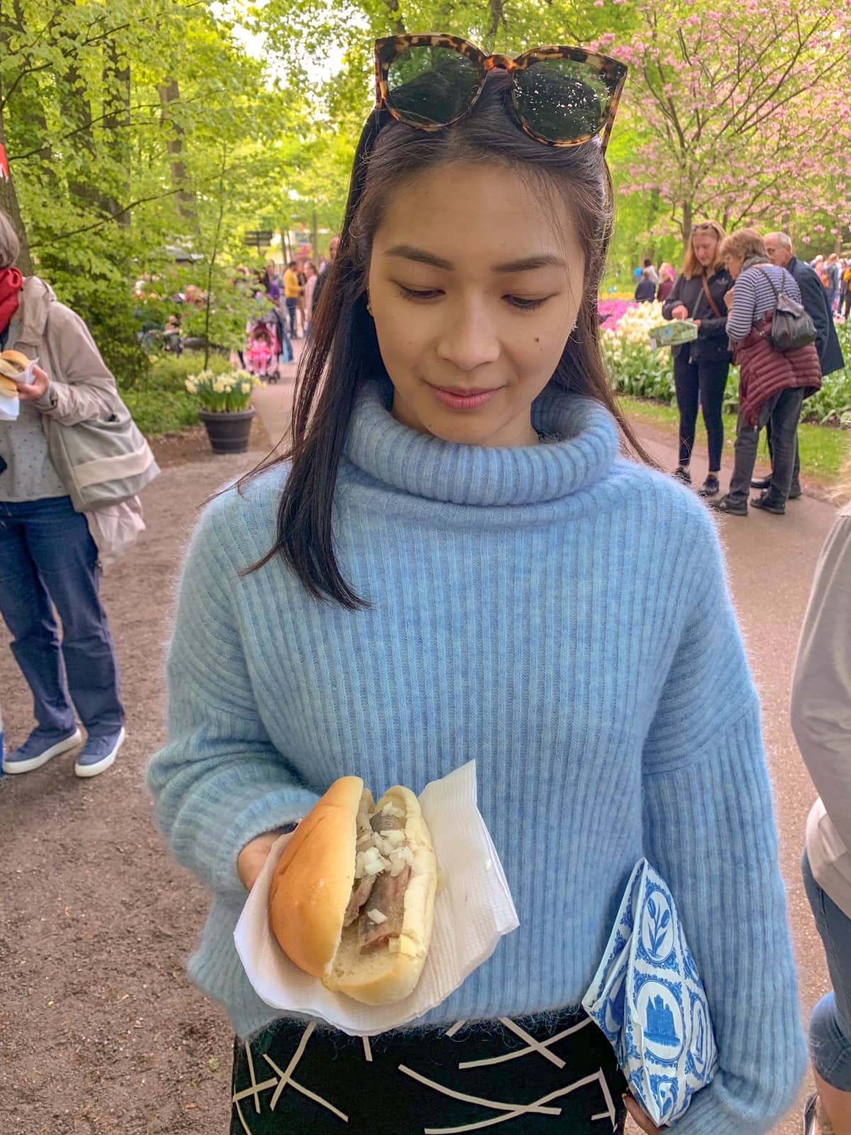 A woman wearing a light blue sweater, holding a napkin with a bun filled with raw herring and onion. She is looking down slightly at the bun with a small smile