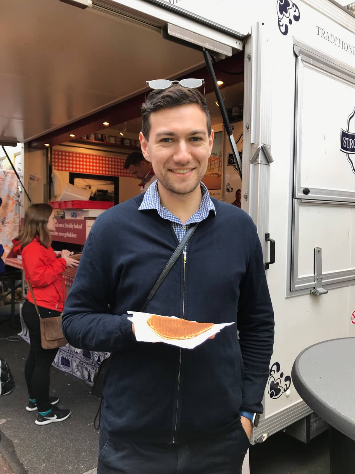 A man in a blue jacket smiling; he’s holding a round flat Dutch stroopwaffel on a napkin