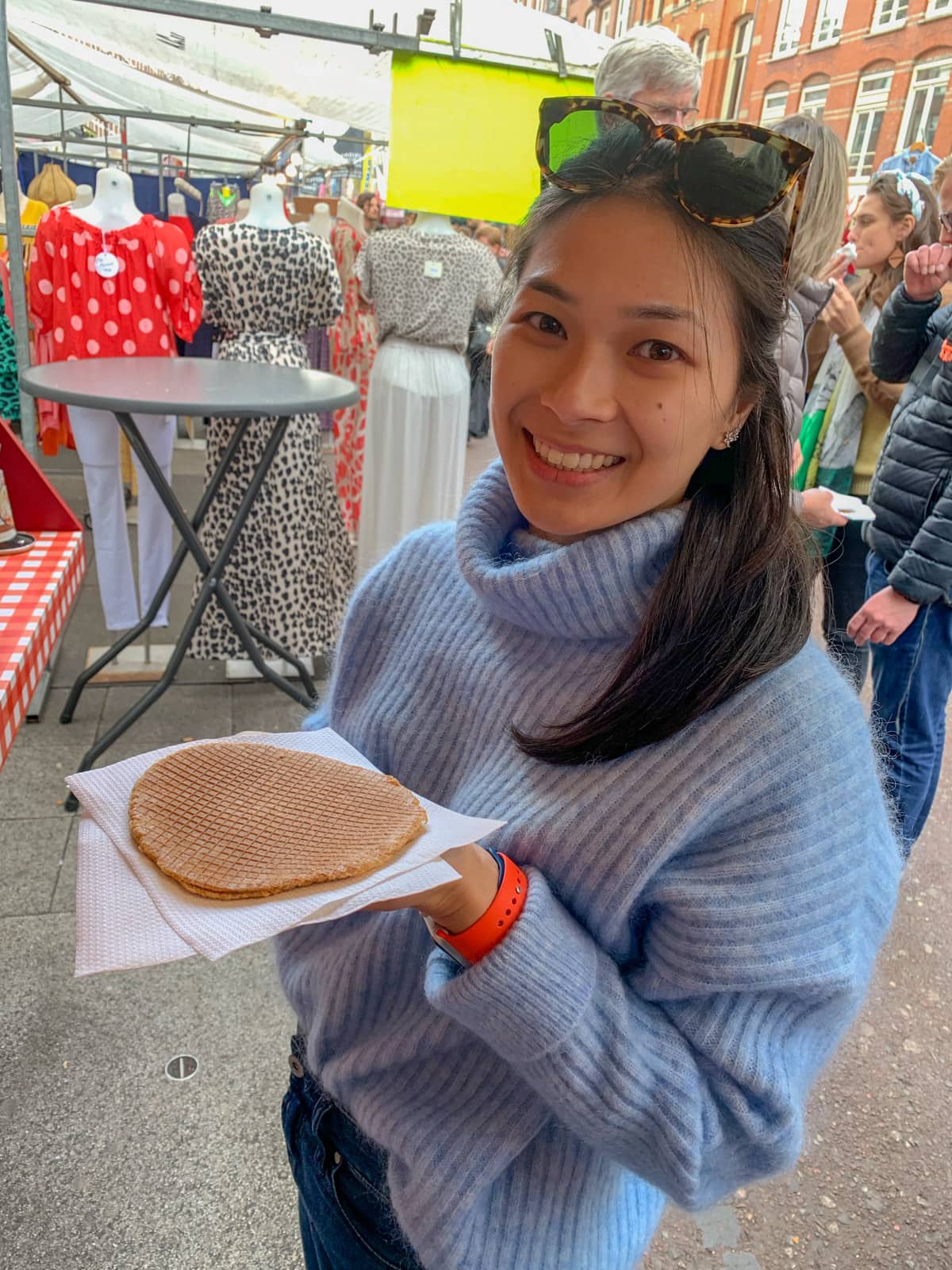 A woman in a light blue sweater smoking; she’s holding a round flat Dutch stroopwaffel on a napkin