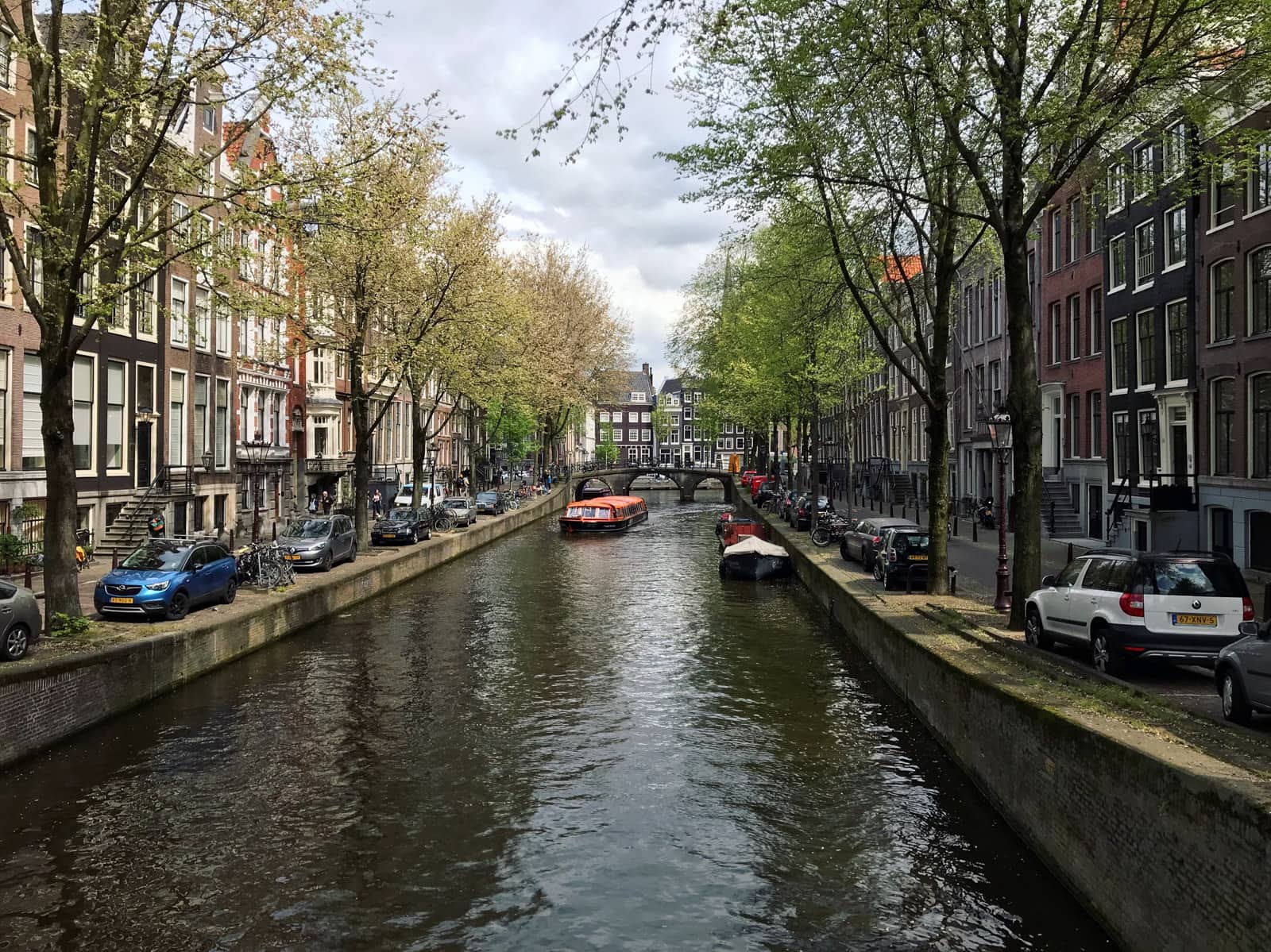 A view of a canal in Amsterdam, seen from a perpendicular bridge. There are a couple of canal boats and there are some trees visible in frame
