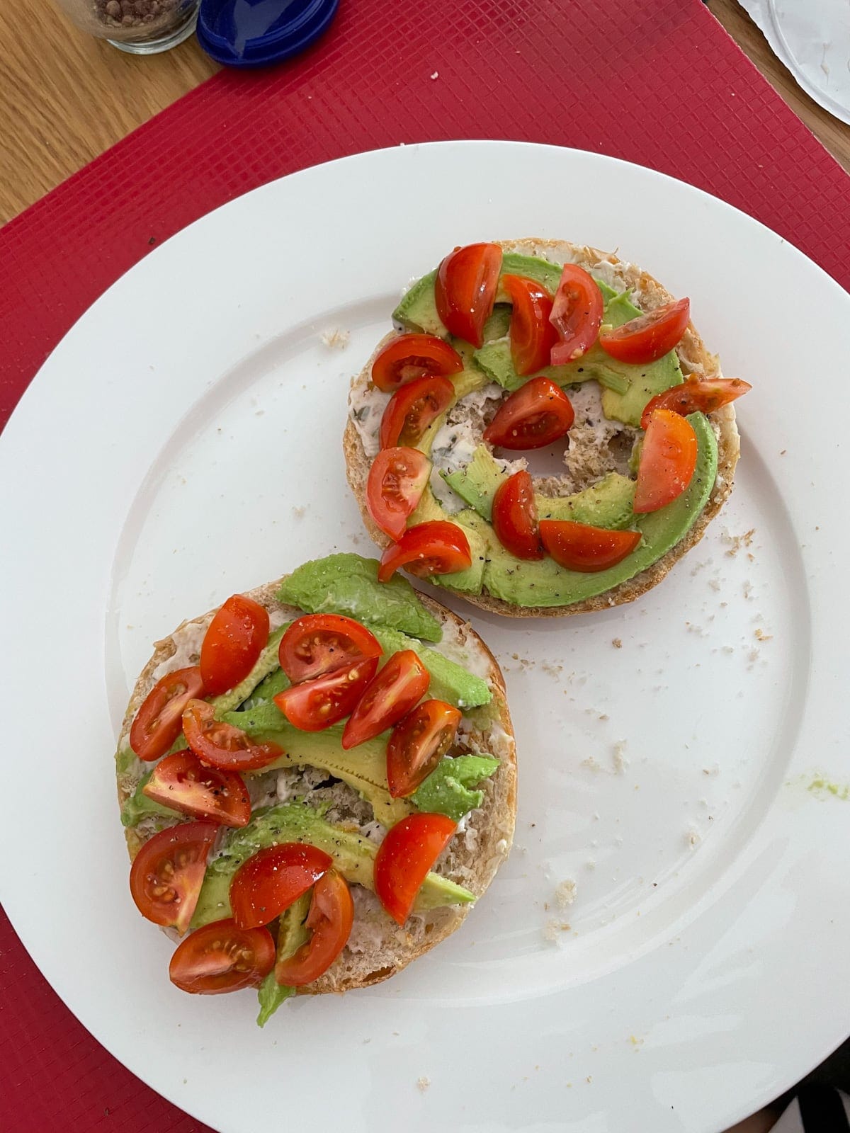 A white plate with an open bagel on it, served with sliced avocado and wedges of cherry tomato