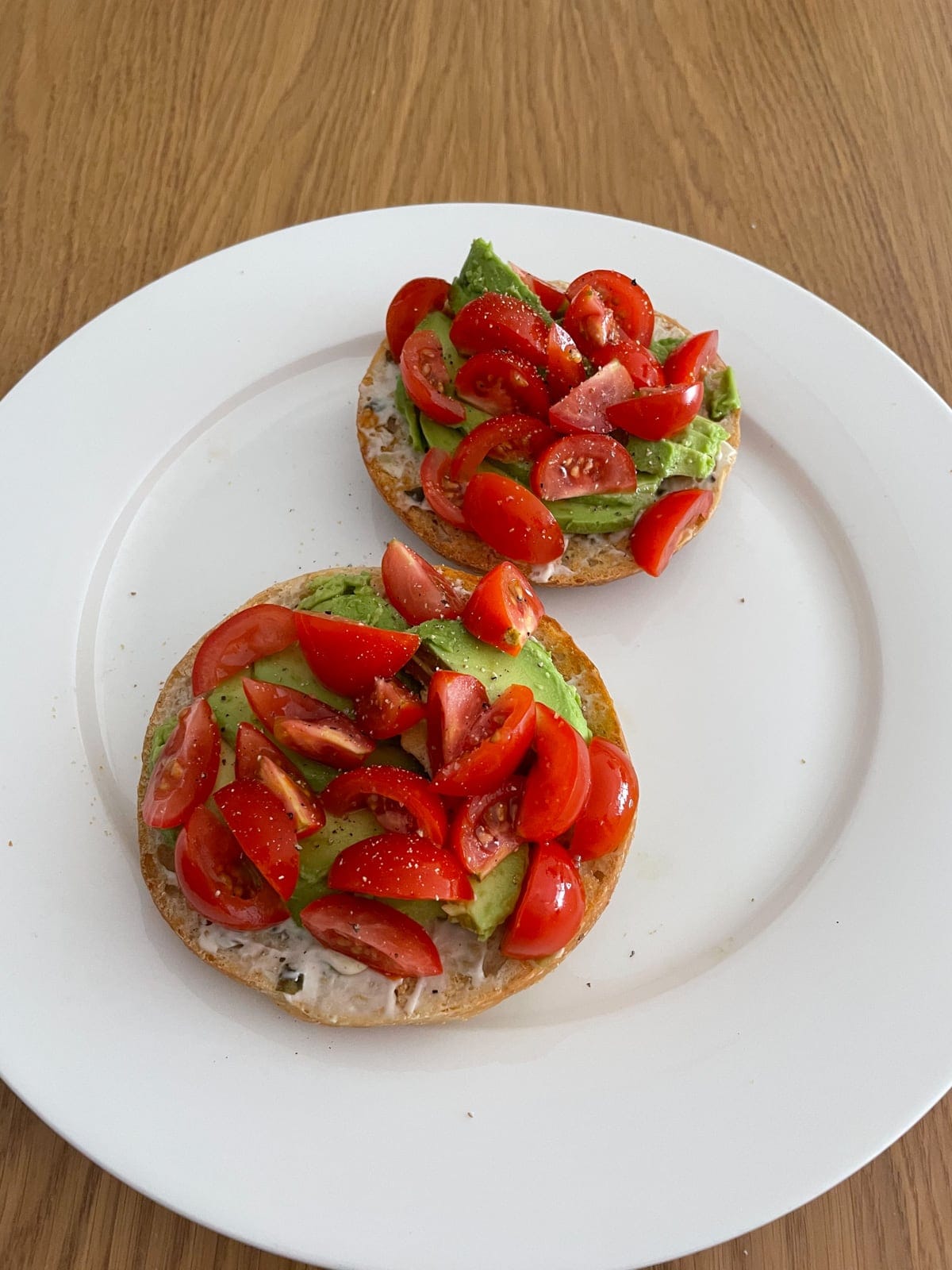 A white plate with an open bagel served on top, with sliced avocado and wedges of cherry tomatoes