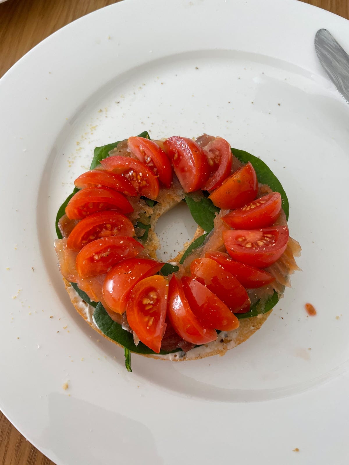 An open bagel served on a white plate, topped with baby spinach leaves, and wedges of cherry tomato arraigned on top neatly like a wheel