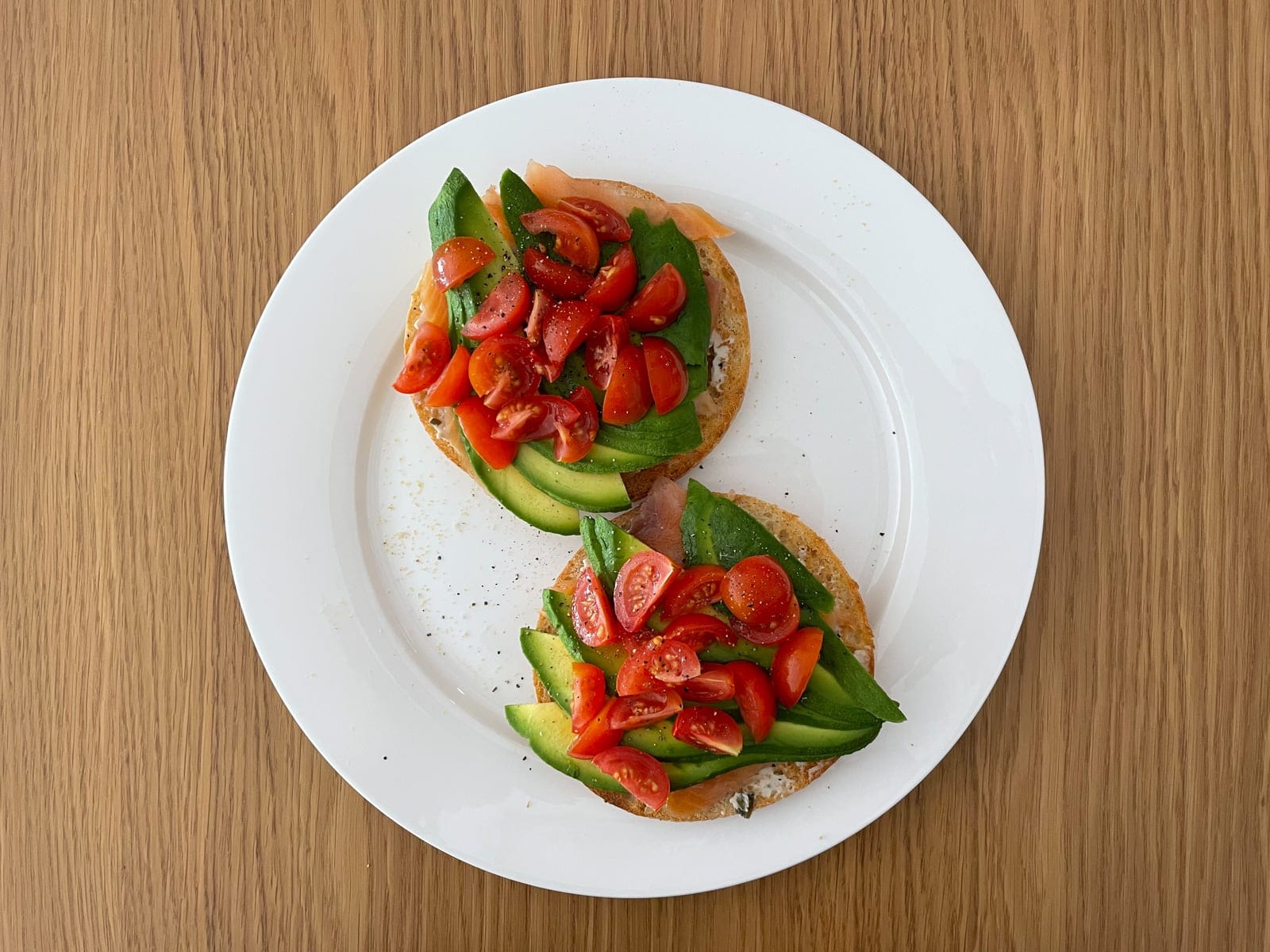 A white plate with an open bagel served with smoked salmon, sliced avocado and a handful of wedges of sliced tomato