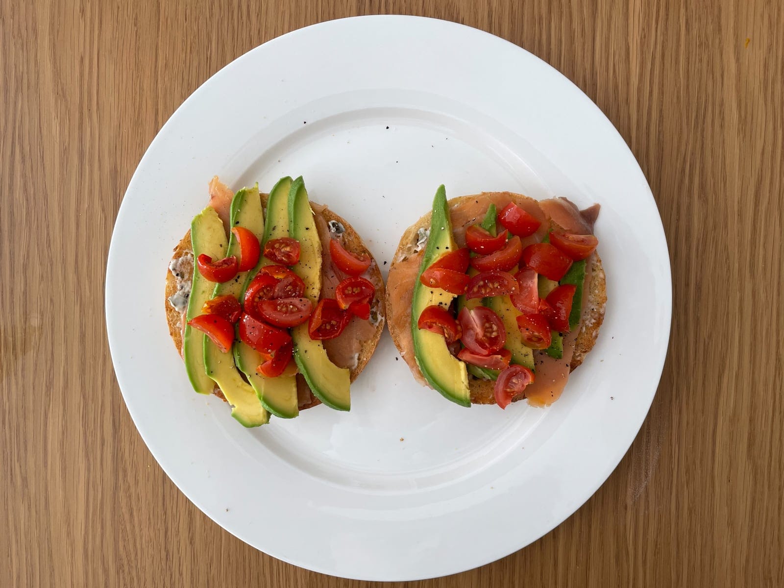 A white plate served with an open bagel of smoked salmon, sliced avocado and wedges of cherry tomatoes