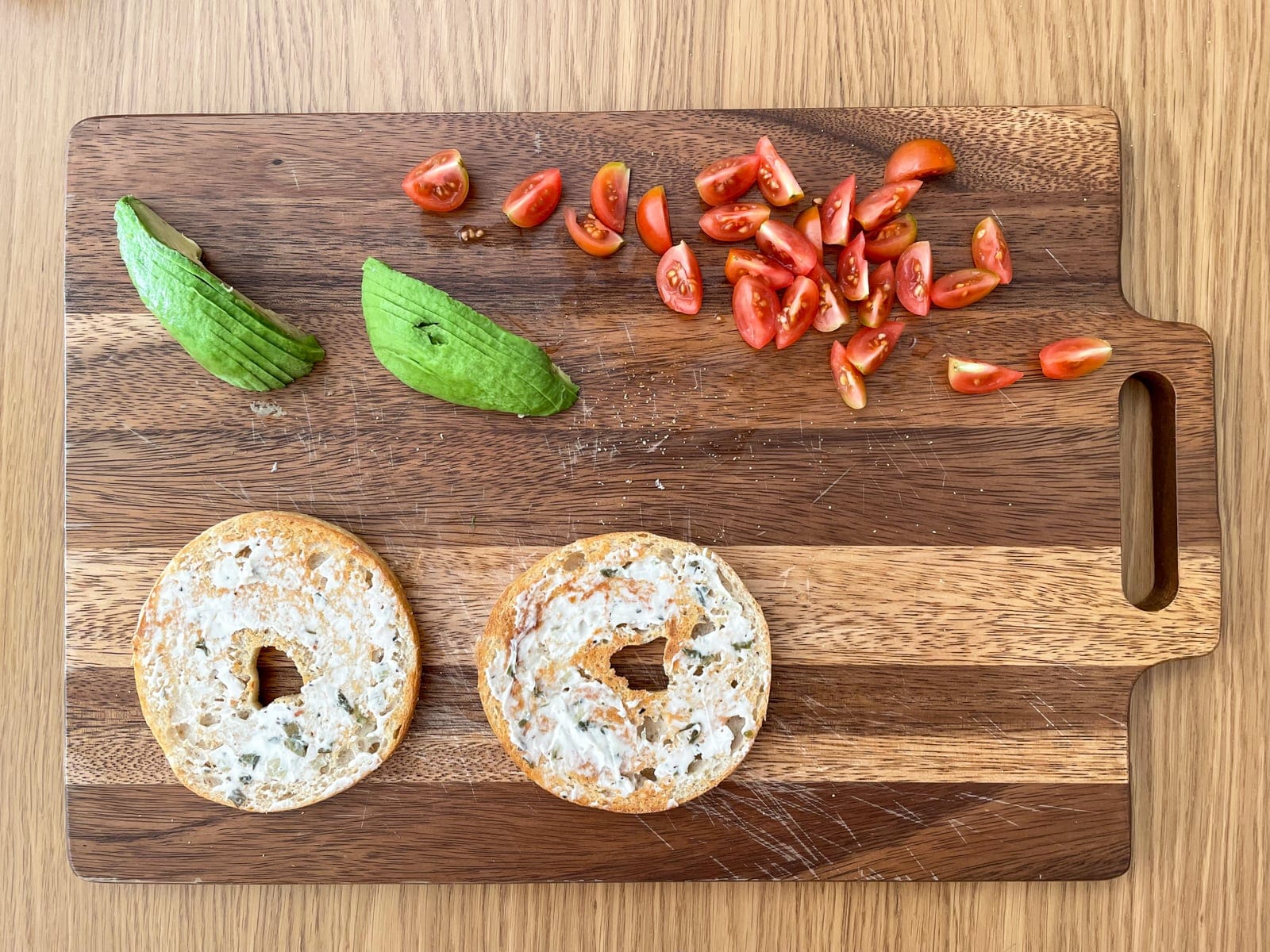 A wooden chopping board laid out with sliced cherry tomatoes and sliced avocado, and an open bagel with cream cheese spread