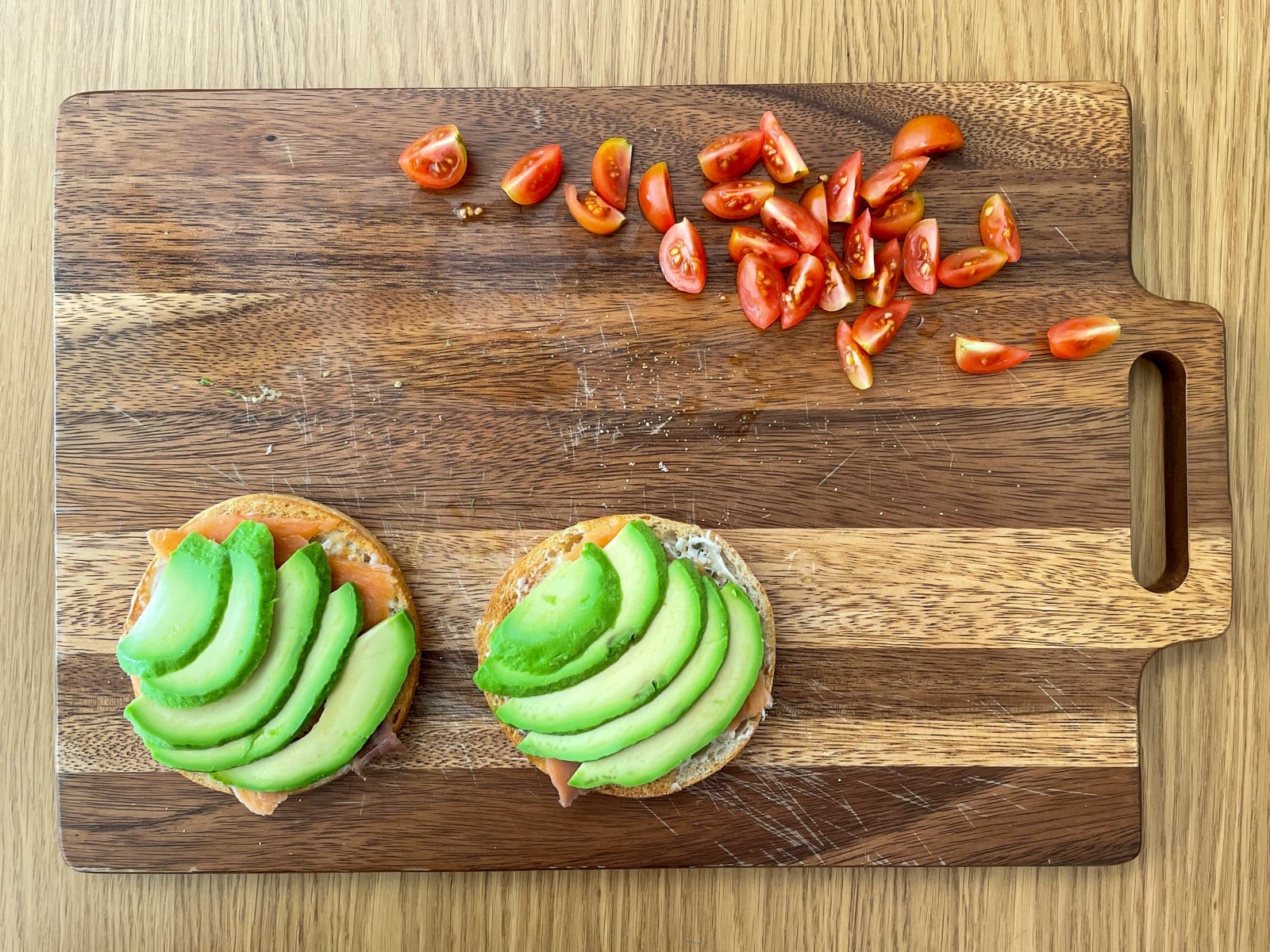 A wooden chopping board laid out with sliced cherry tomatoes and an open bagel with sliced avocado fanned out on top.