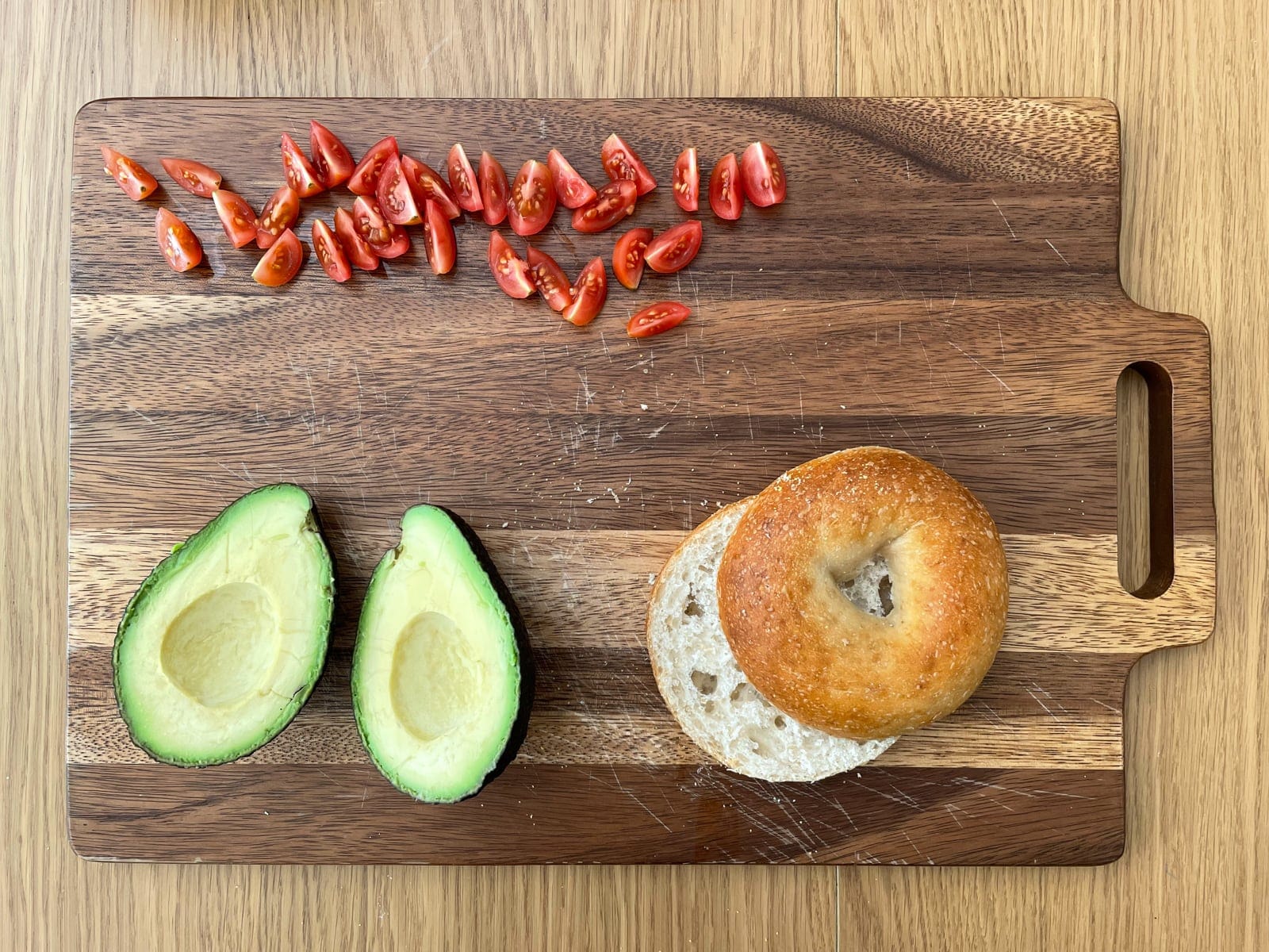 A wooden chopping board with wedges of sliced cherry tomato, a cut open avocado, and a bagel cut in half