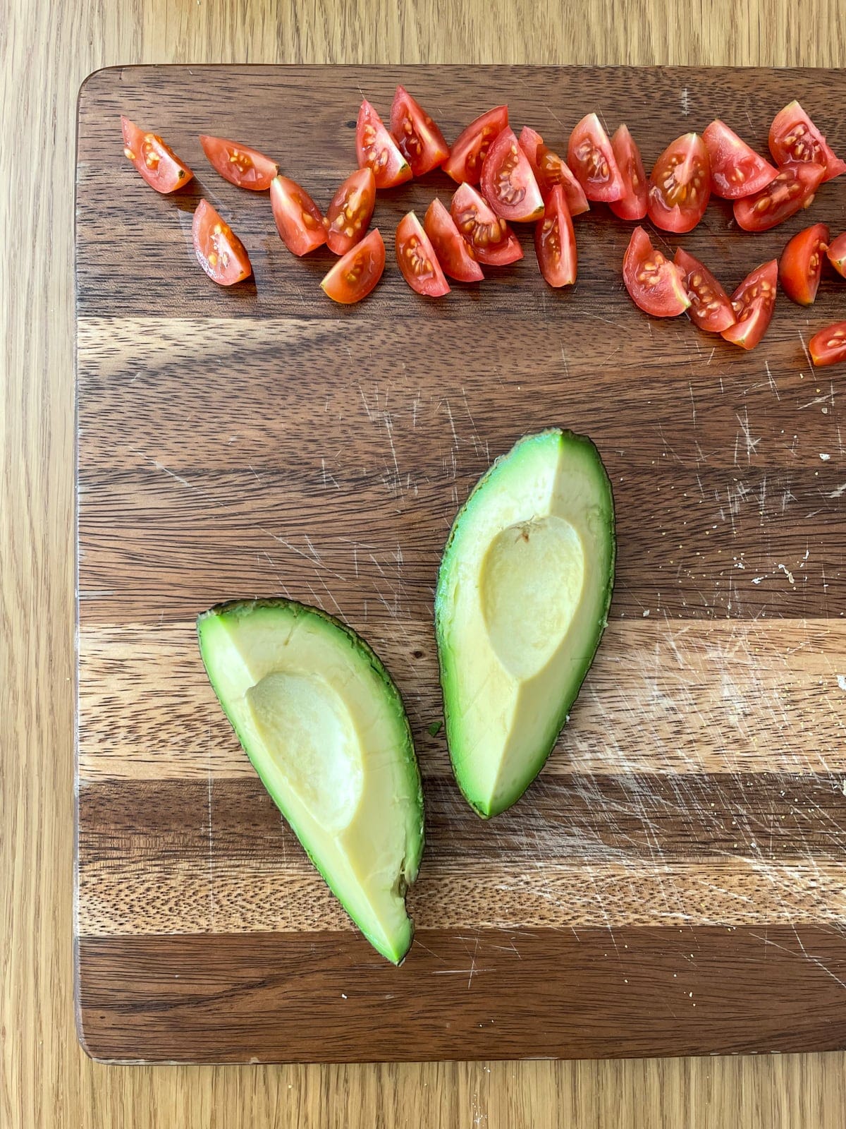 A close-up of two avocado quarters, unpeeled, on a wooden chopping board alongside some sliced cherry tomatoes