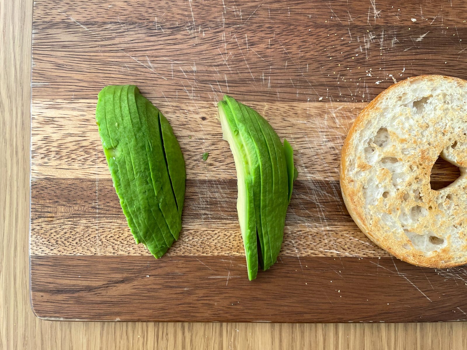 A close-up photo of two peeled and sliced avocado quarters, sitting on a wooden chopping board next to an open half of a bagel