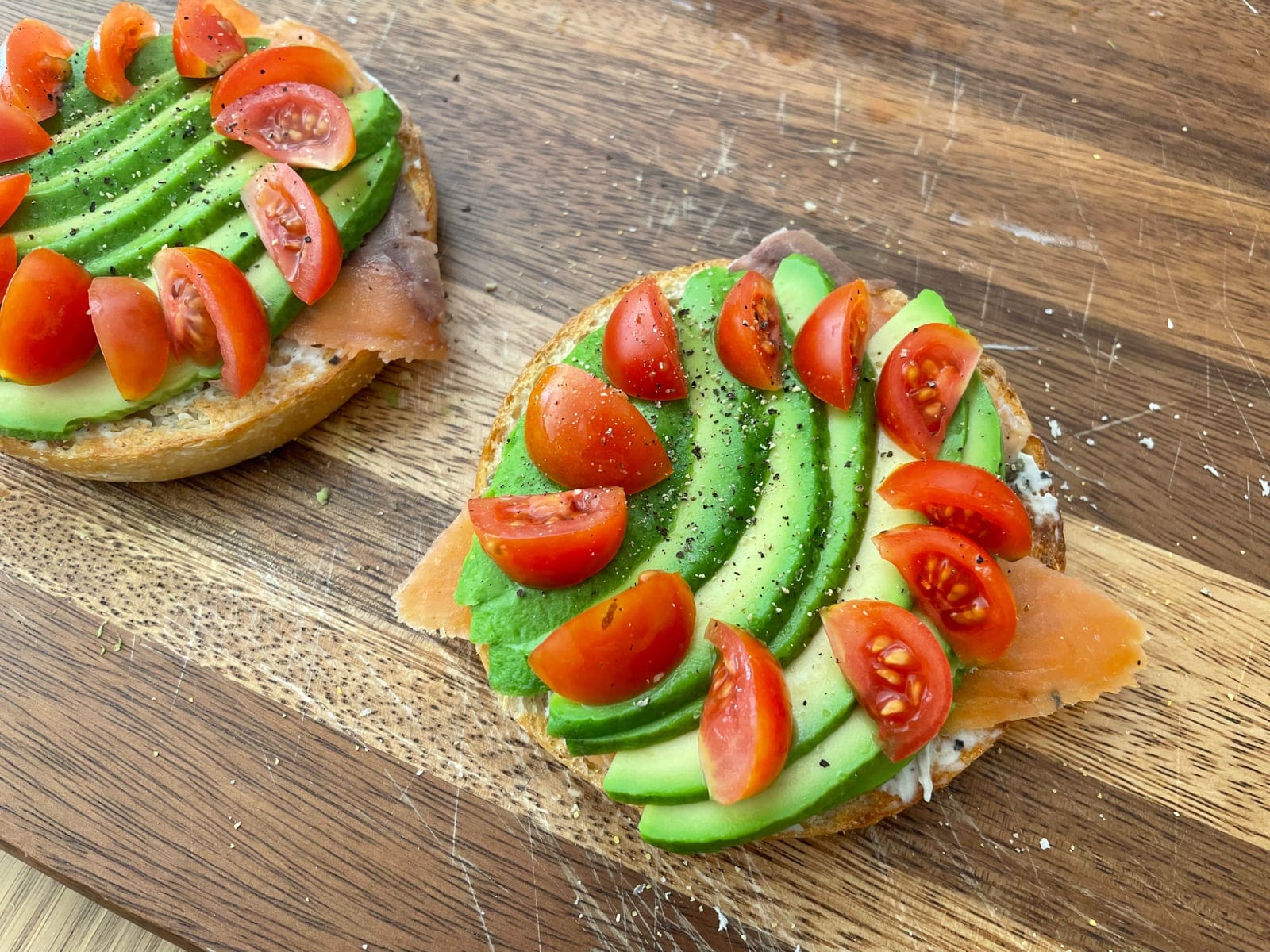 A close-up of an open bagel assembled with smoked salmon, sliced avocado and wedges of cherry tomato arranged in a circle