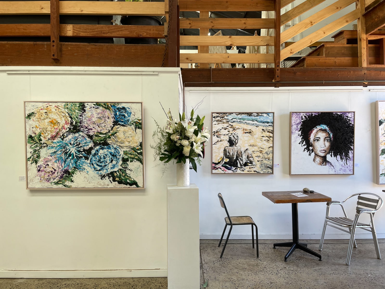 The inside of an art gallery during the day. There are some pieces of art on the walls, and a small table and chairs in the corner. There is a second level to the building which has a wooden railing