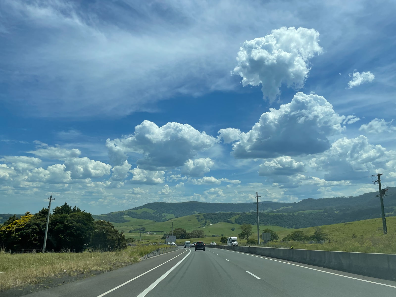 A view of a stretch of road during the day. The sky has some clouds in it. The road is a motorway with some cars on the road
