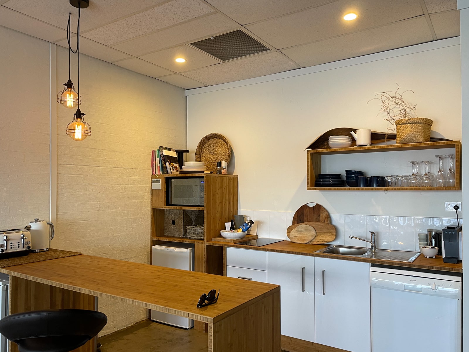 A kitchen with a wooden high table, wooden shelving, white whitegoods and cupboards, and some lighting hanging from the ceiling