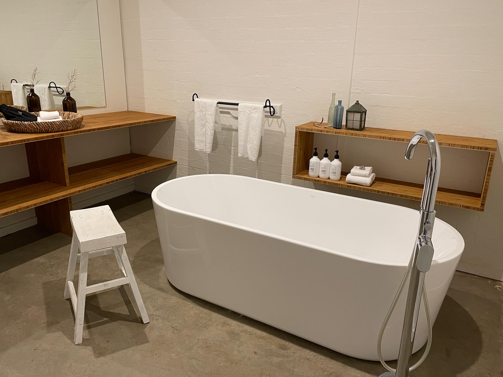 A big white bathtub placed on the ground of a bathroom. A white painted wooden stool sits next to it. There is brown wooden shelving against the walls.