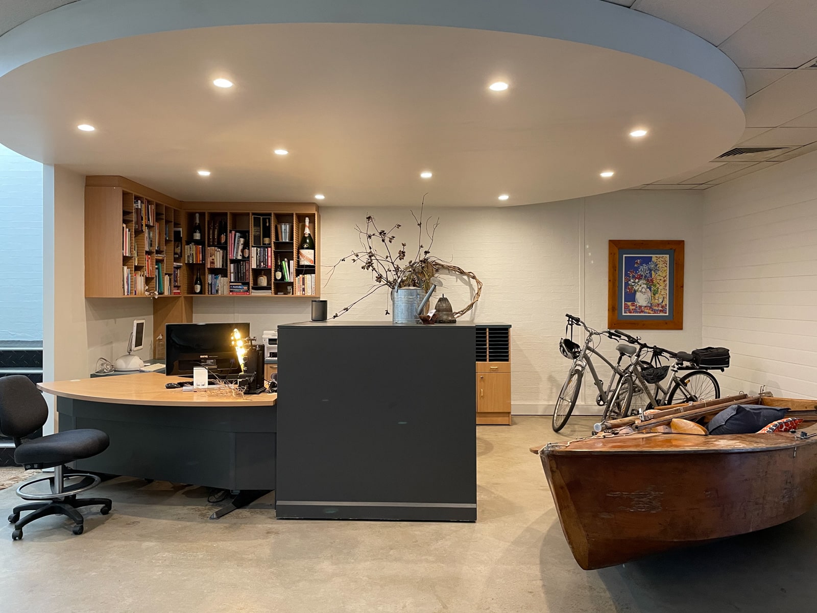 The common area of a hotel, resembling a cosy office. There is a desk with a computer and some shelving on the walls, attached close to the ceiling. To the side is a wooden boat, as decoration, with some cushions in it. Two bicycles sit in the corner behind the boat.