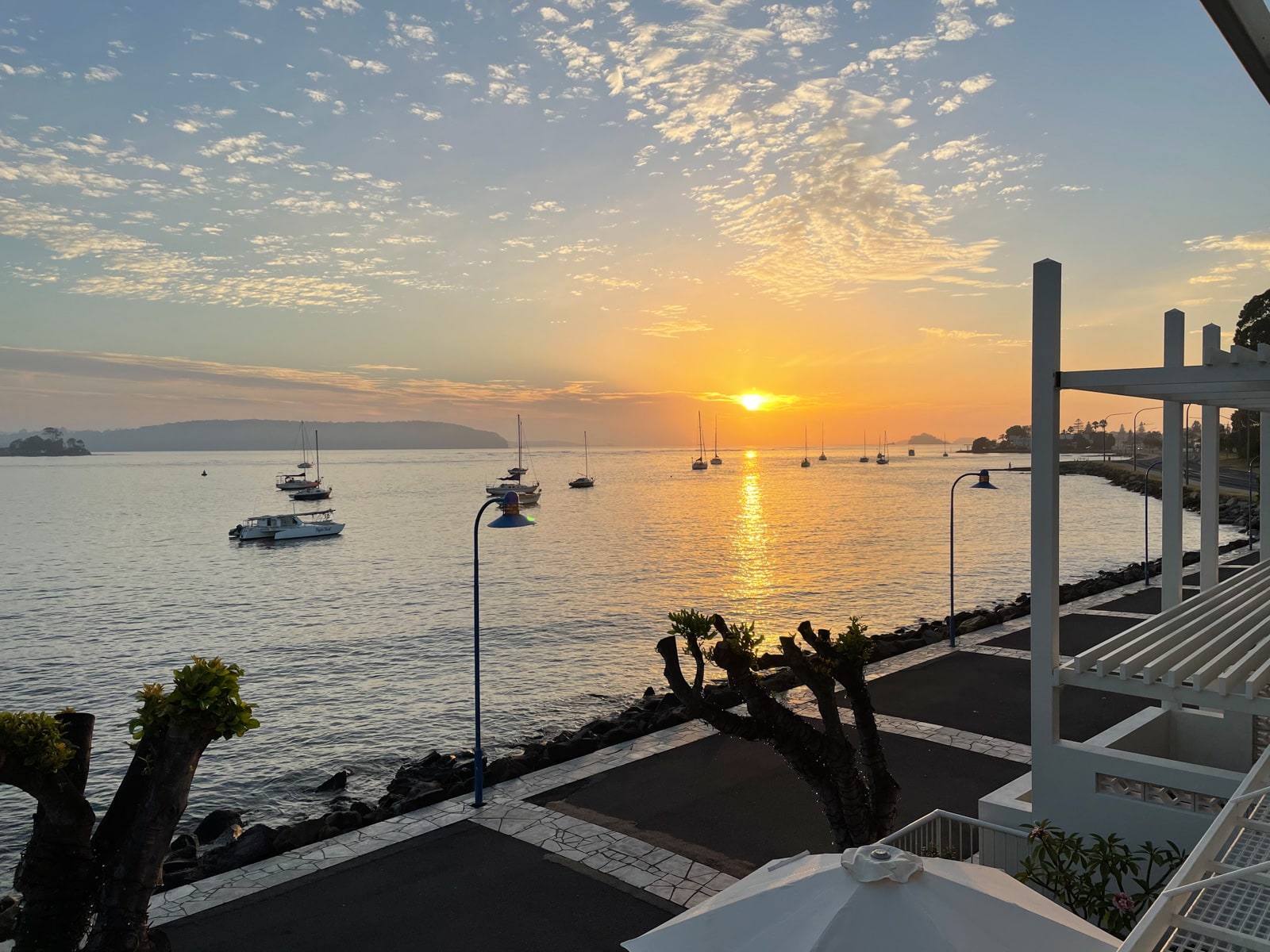 A view from a second floor balcony, of the sunrise on the water. The promenade can be seen down below. There are streaks of clouds across the sky