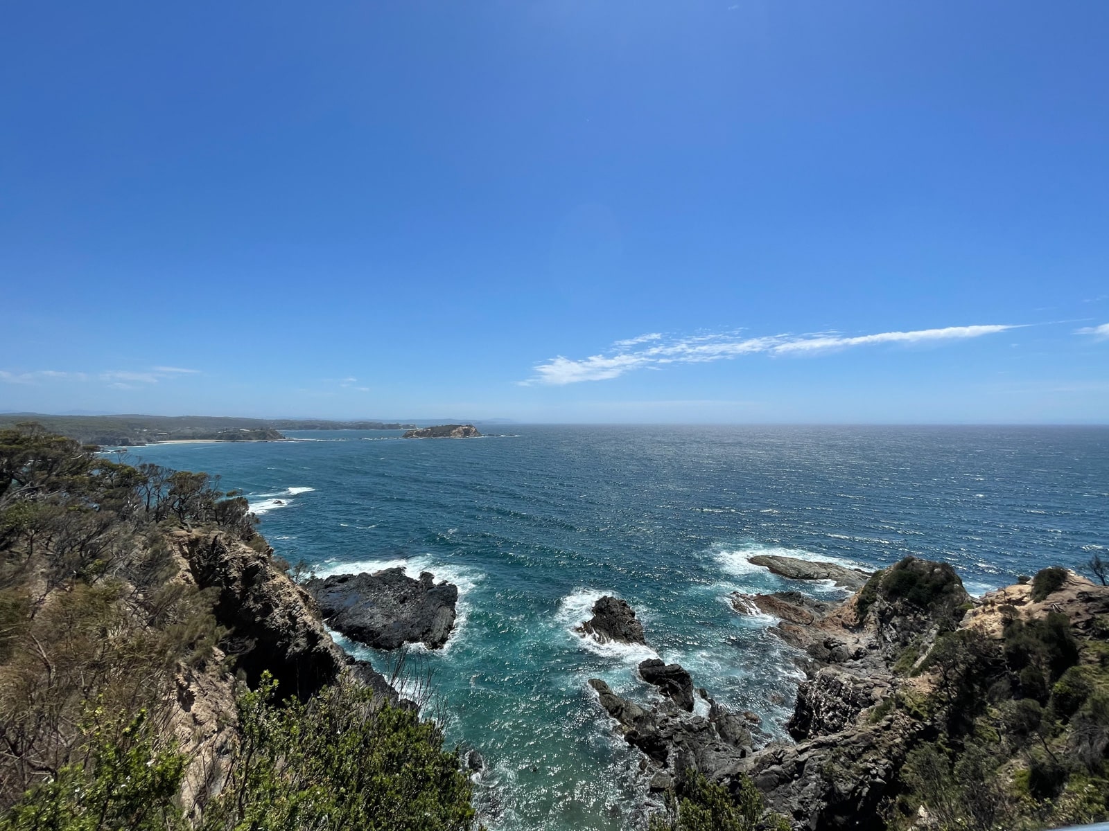A view of the ocean from a viewing platform, with dark rock formations in the foreground. It is a sunny day with an almost cloudless sky