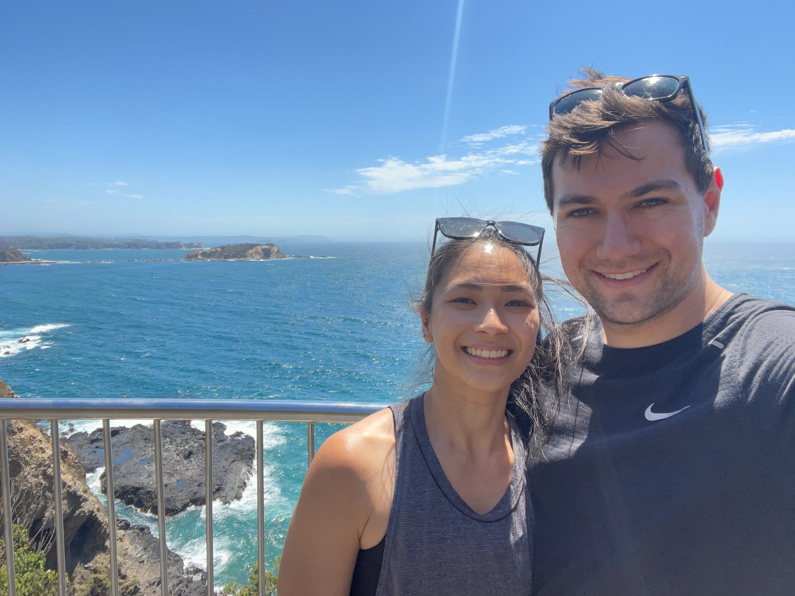 A selfie of a man and woman, both with sunglasses on their head, and a view of the ocean in the background. It is a sunny day