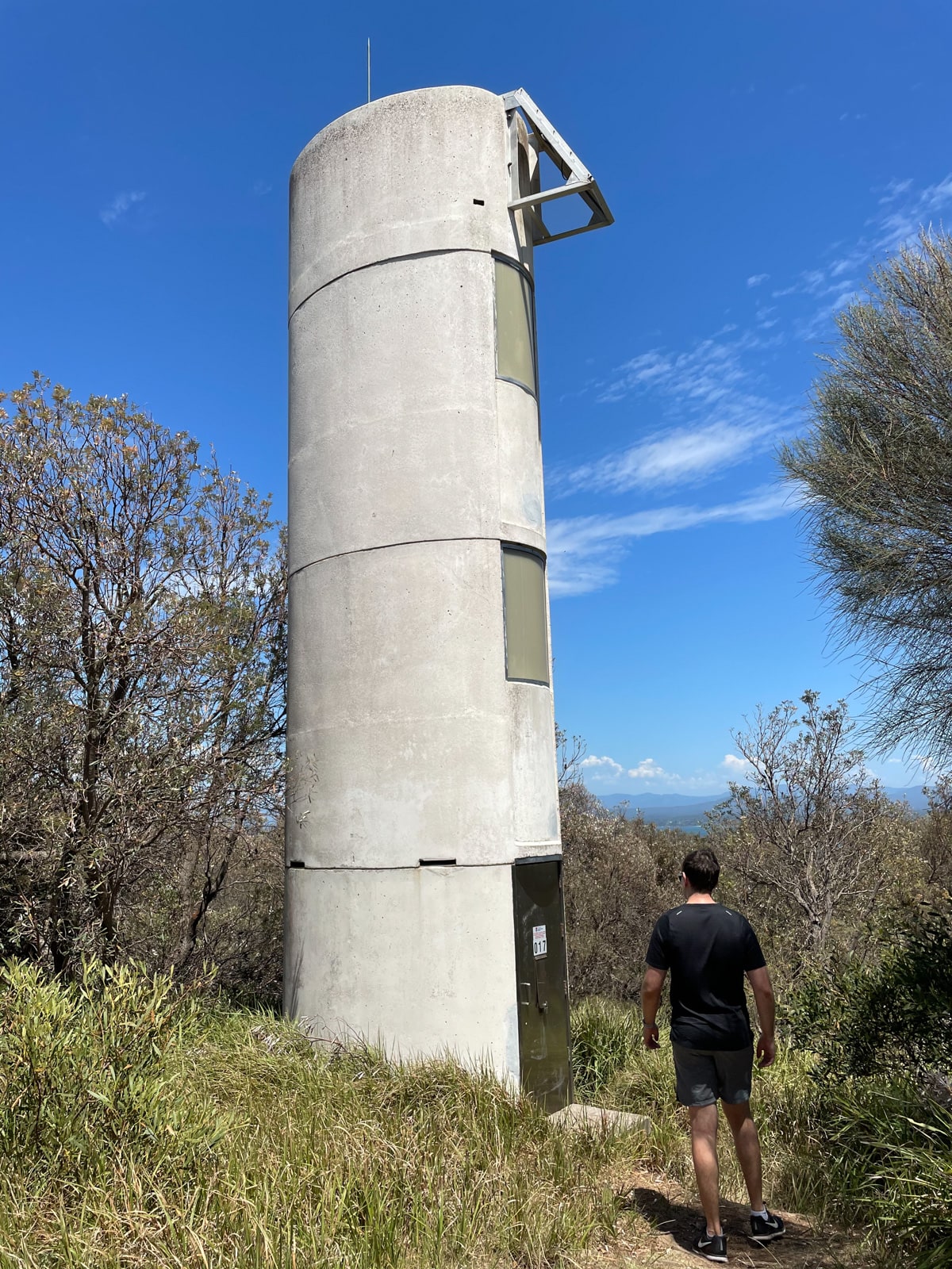 A man walking towards a lightstation that is several storeys tall. It appears to be in the middle of bushland