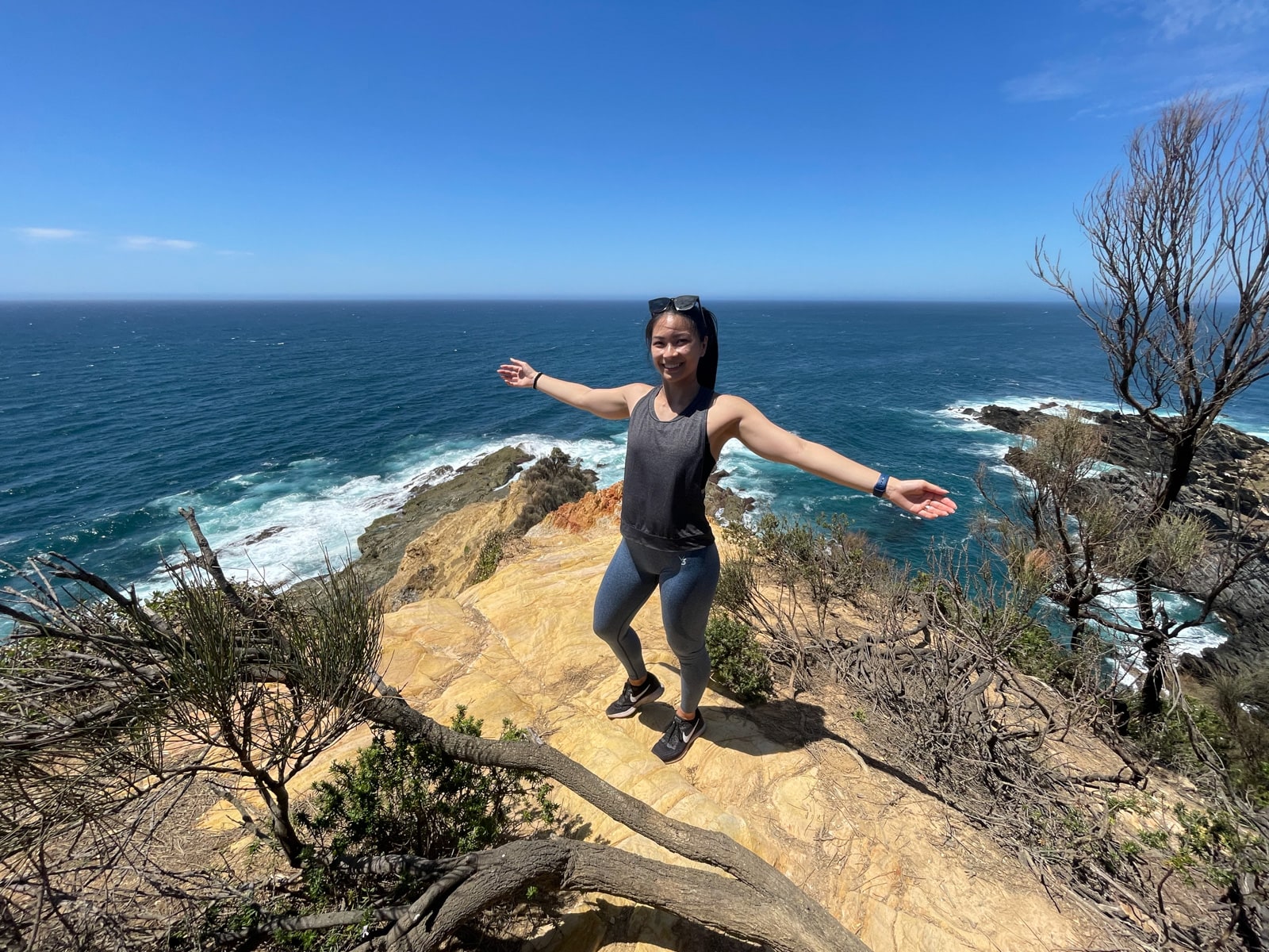 A woman with outstretched arms, dressed in activewear. She is standing on an earthy yellow cliff face and smiling, with a great view of the deep blue ocean behind her. In the foreground, on the cliff, are barren-looking trees with only branches left