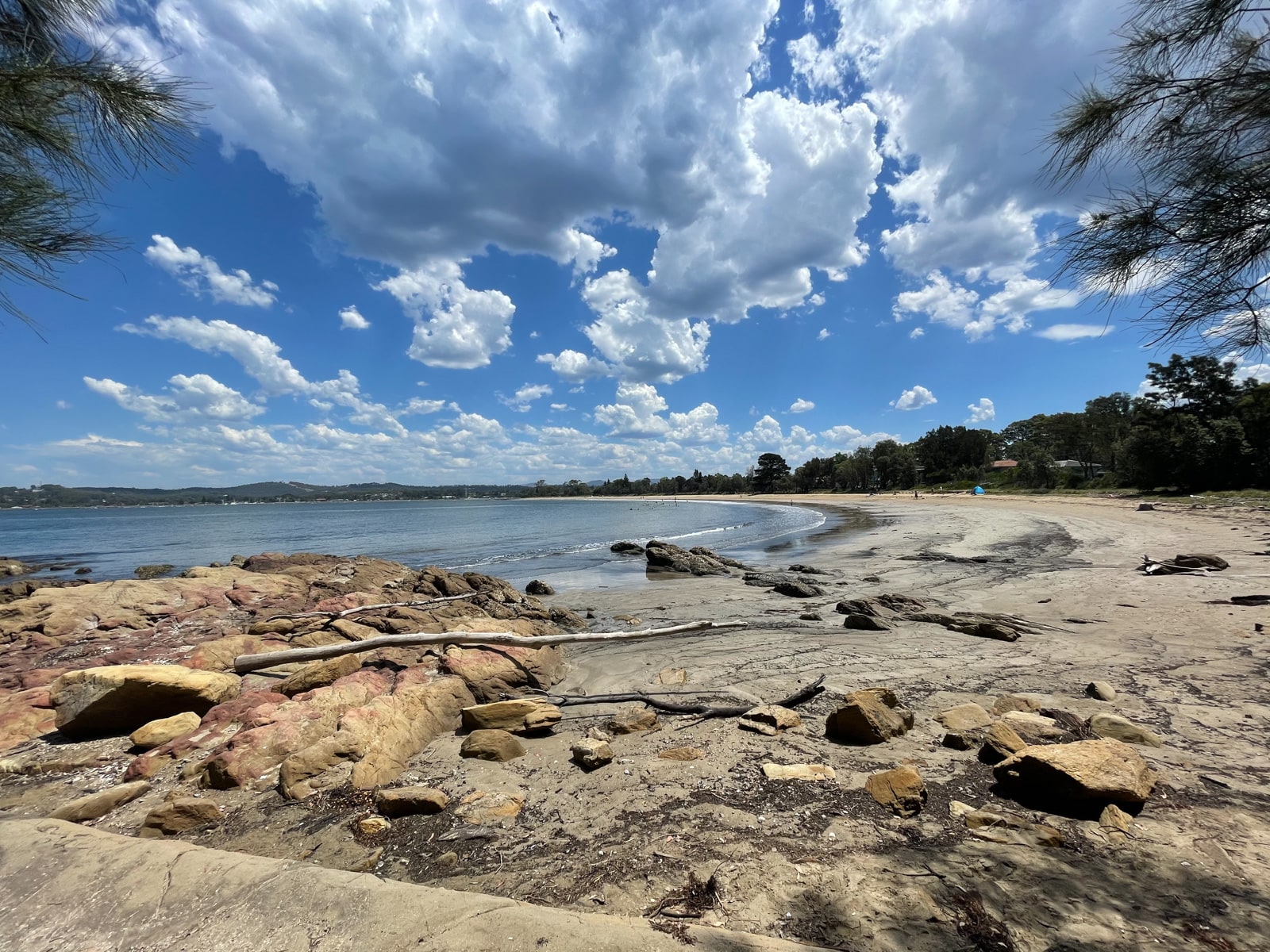 A dirt beach with large rocks on the sand surface. The water is at very low tide and the beach curves around into the distance. The blue sky is covered in clouds