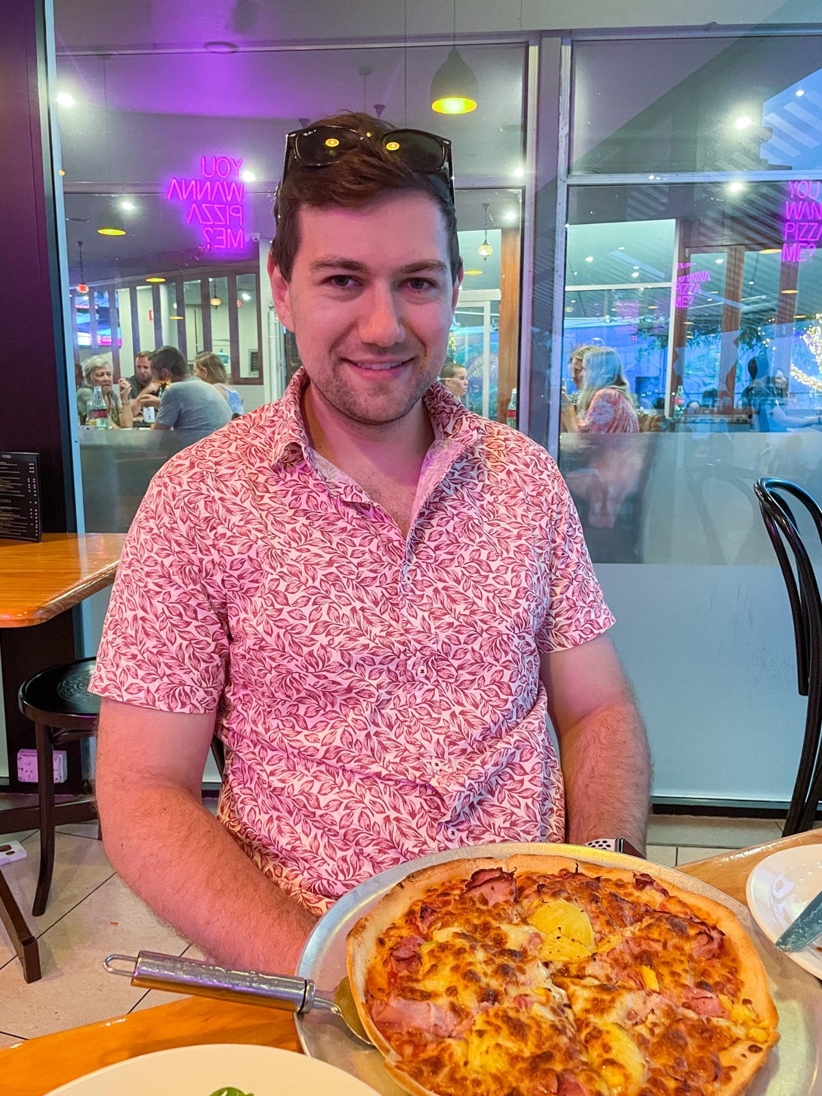 A man with dark brown hair, in a red summery patterned shirt, sitting at a table with Hawaiian pizza