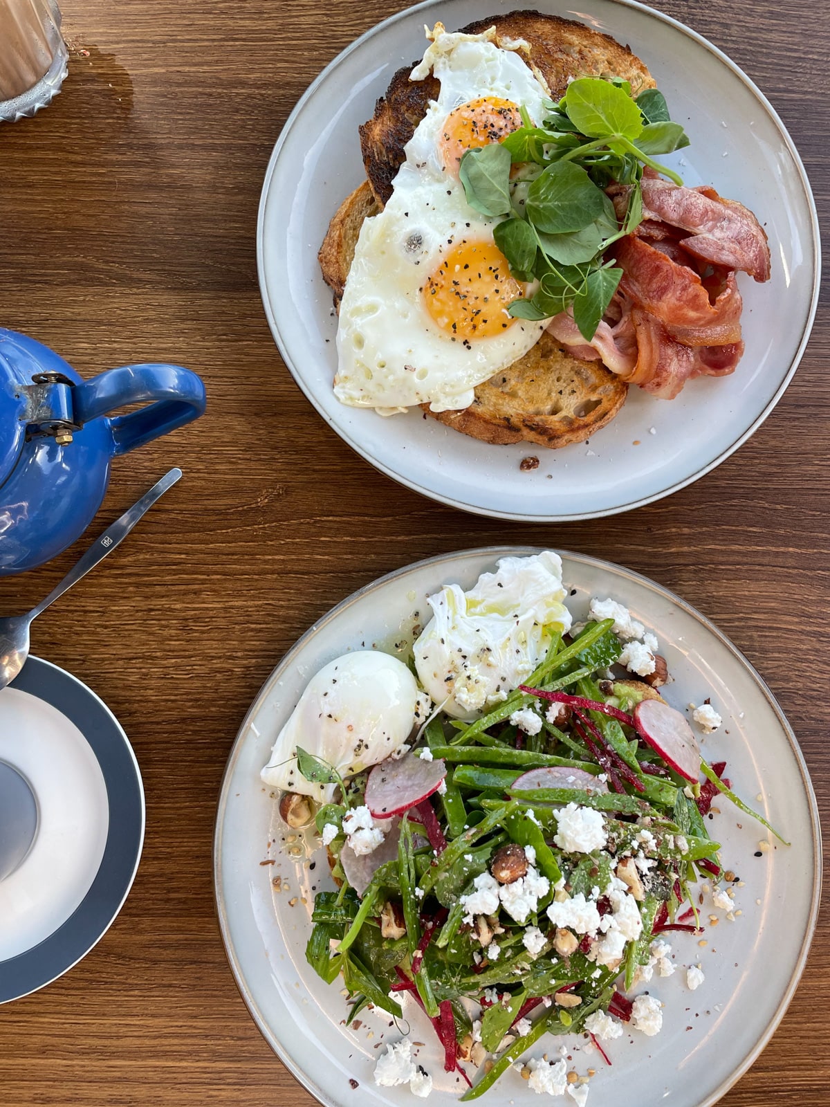A birds-eye view of a wooden table with two plates; one plate is served with greens and avocado on toast with poached eggs and feta; the other is served with two fried eggs on toast with bacon