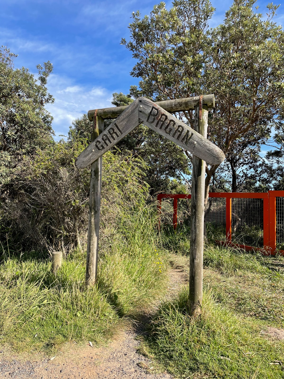 A tall arch, made from wood, over a dirt path, with “Gari bagan” carved into a boomerang-shaped sign on top.