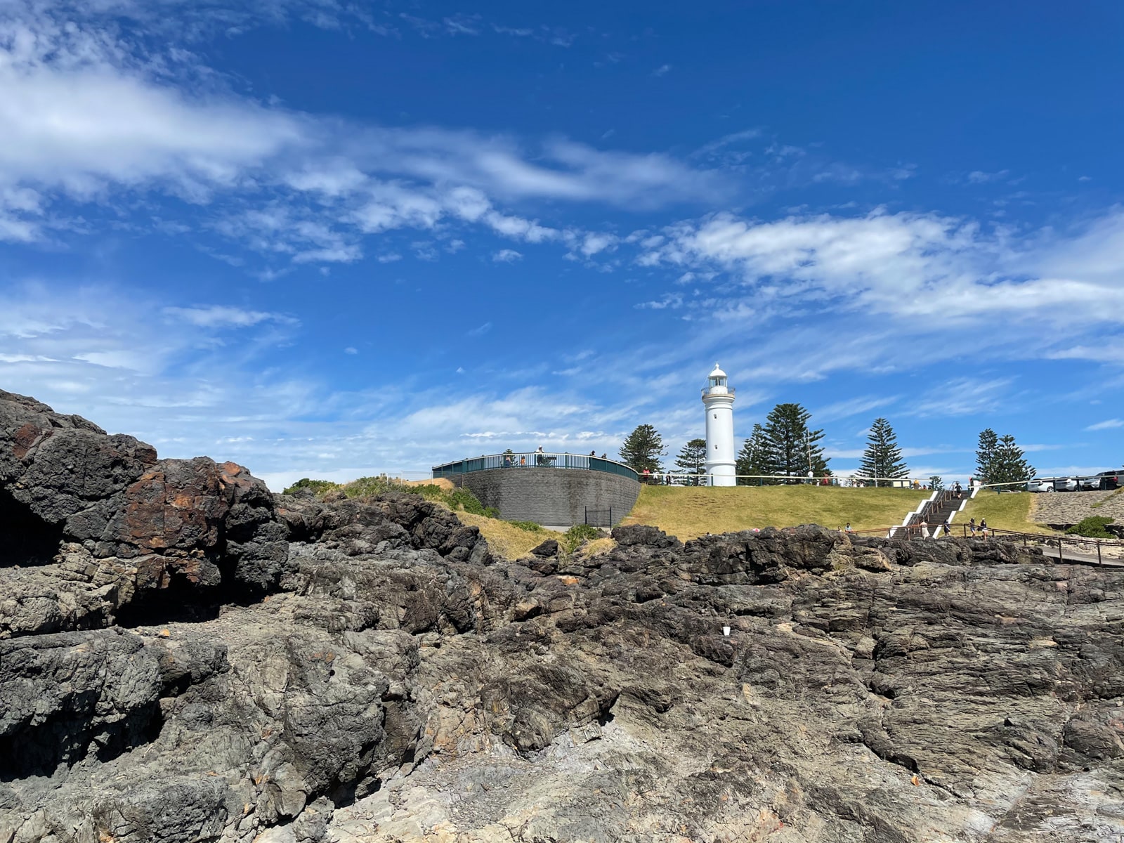 A lighthouse with stairs leading up to it and a viewing platform. In the foreground are big rocks forming an uneven surface. The sky is blue with several clouds