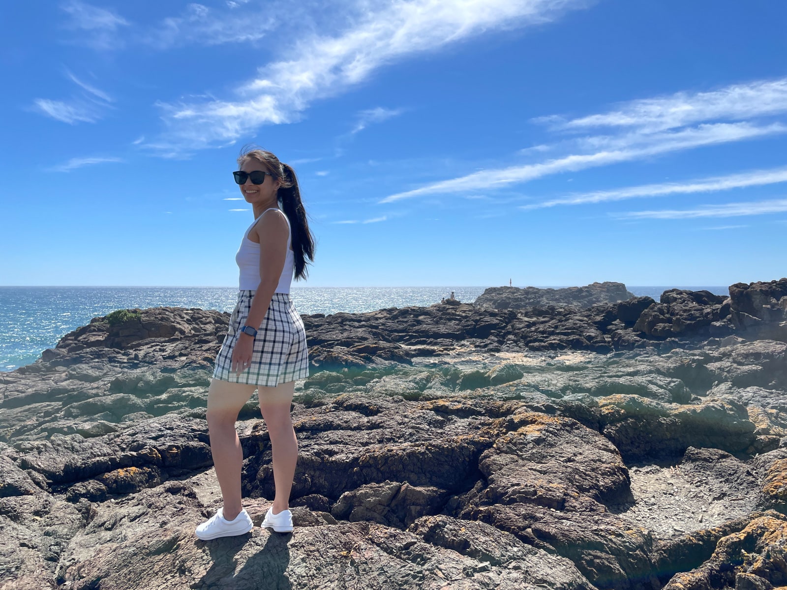 A woman with long dark hair in a ponytail, wearing sunglasses. She is dressed in a white top and checkered shorts and white shoes, and is standing on a big rocky surface with the ocean in the background