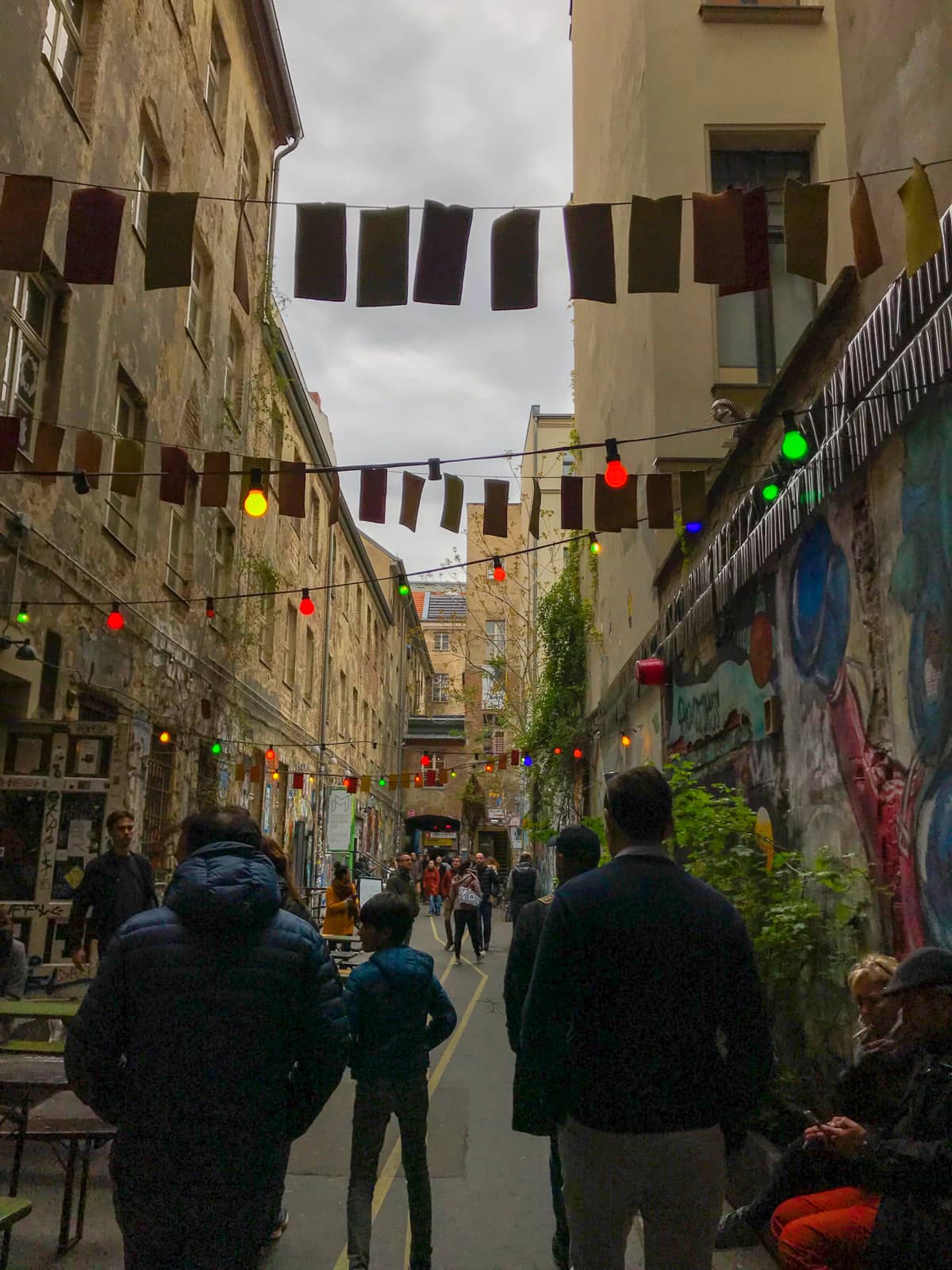 A laneway with coloured lights hanging from wires across the laneway like a canopy, some people walking in the middle of the frame, and street art on the walls of the buildings on either side.